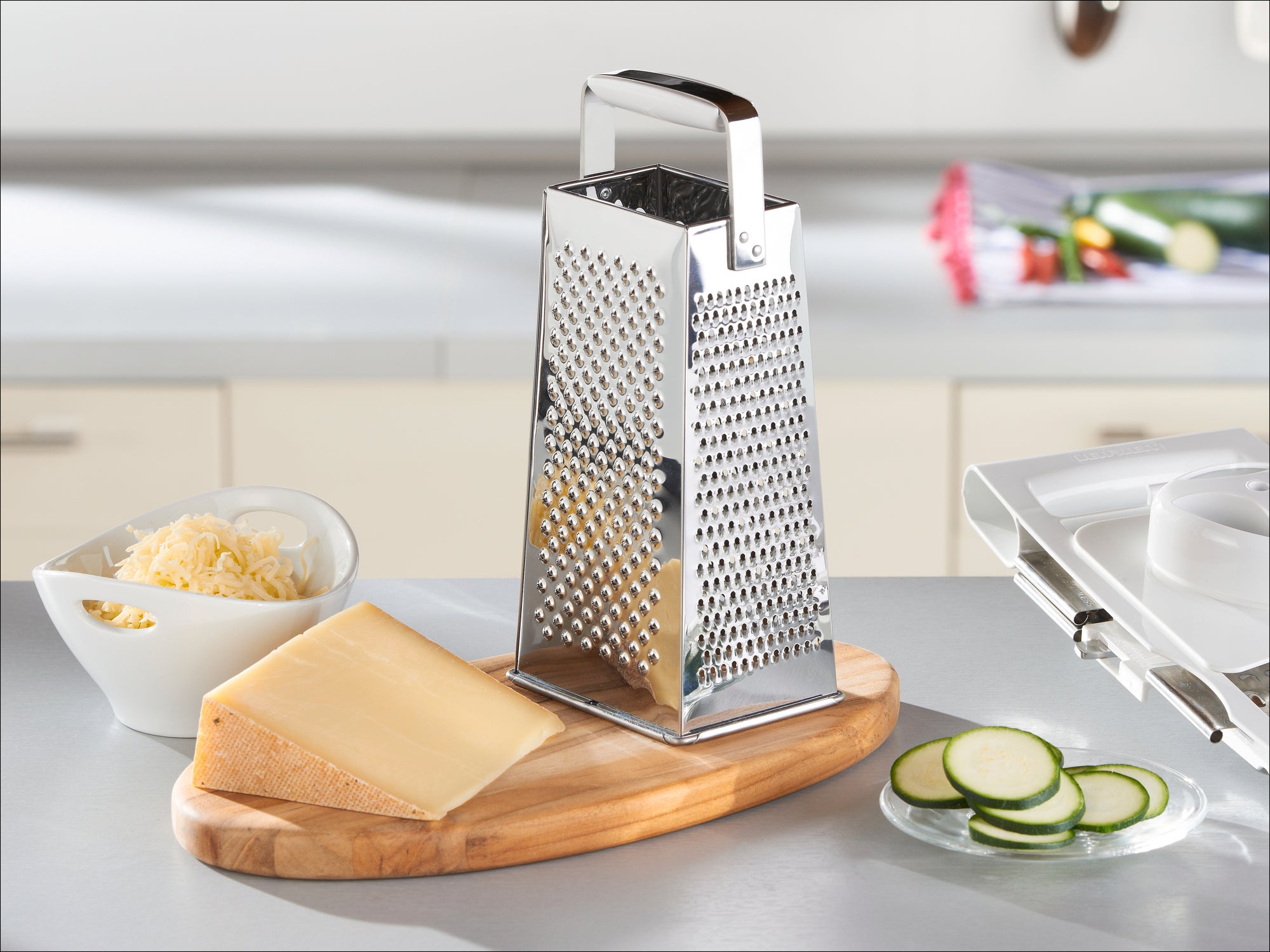 Rechteckige Edelstahlreibe auf einer Küchenarbeitsplatte, umgeben von geriebenem Käse in einer Schüssel, einem Stück Käse und geschnittenen Zucchini auf einem Holzbrett.