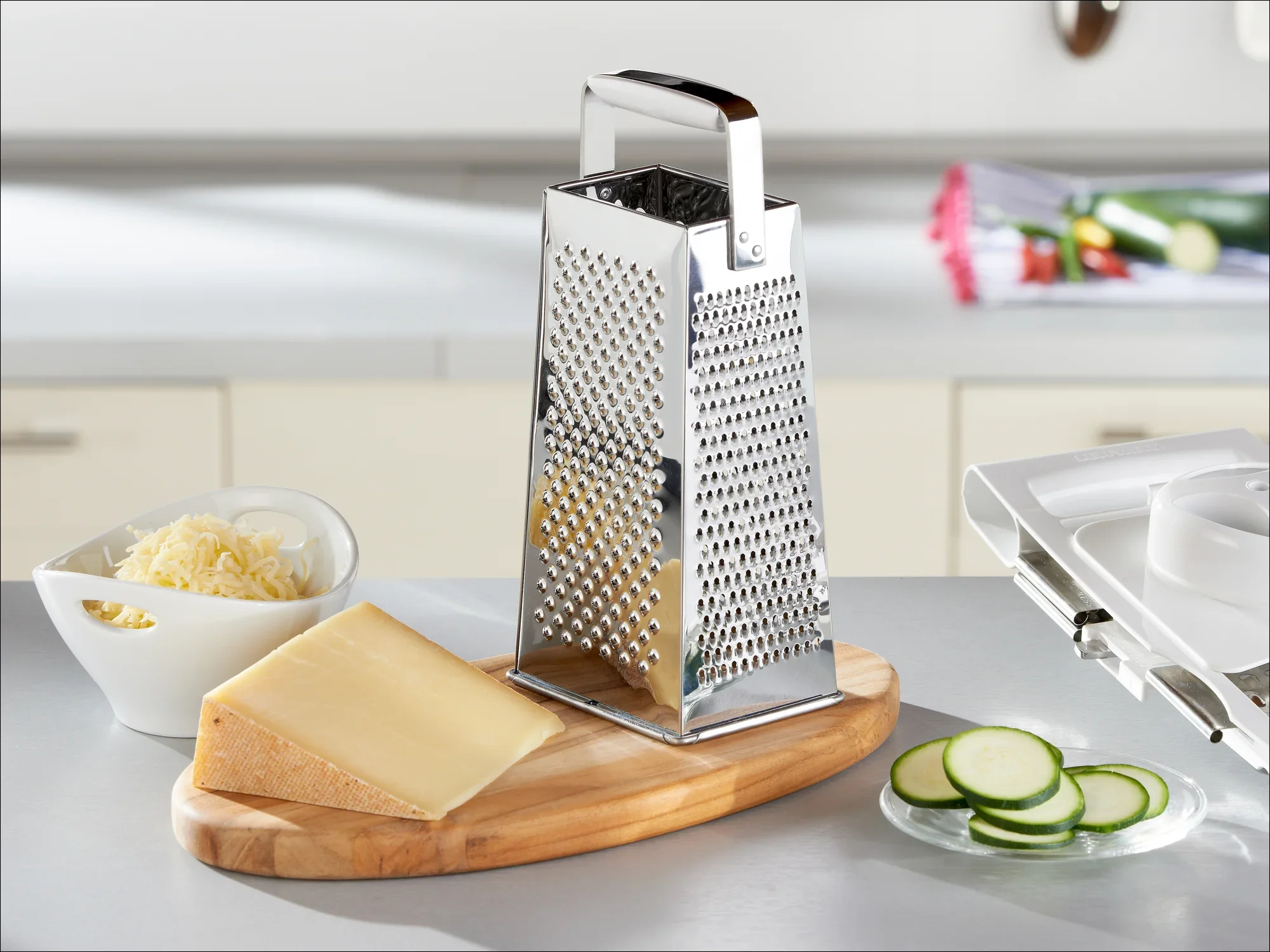 Rechthoekige rasp van roestvrij staal op een keukenblad, omringd door geraspte kaas in een kom, een stuk kaas en gesneden courgettes op een houten plank.