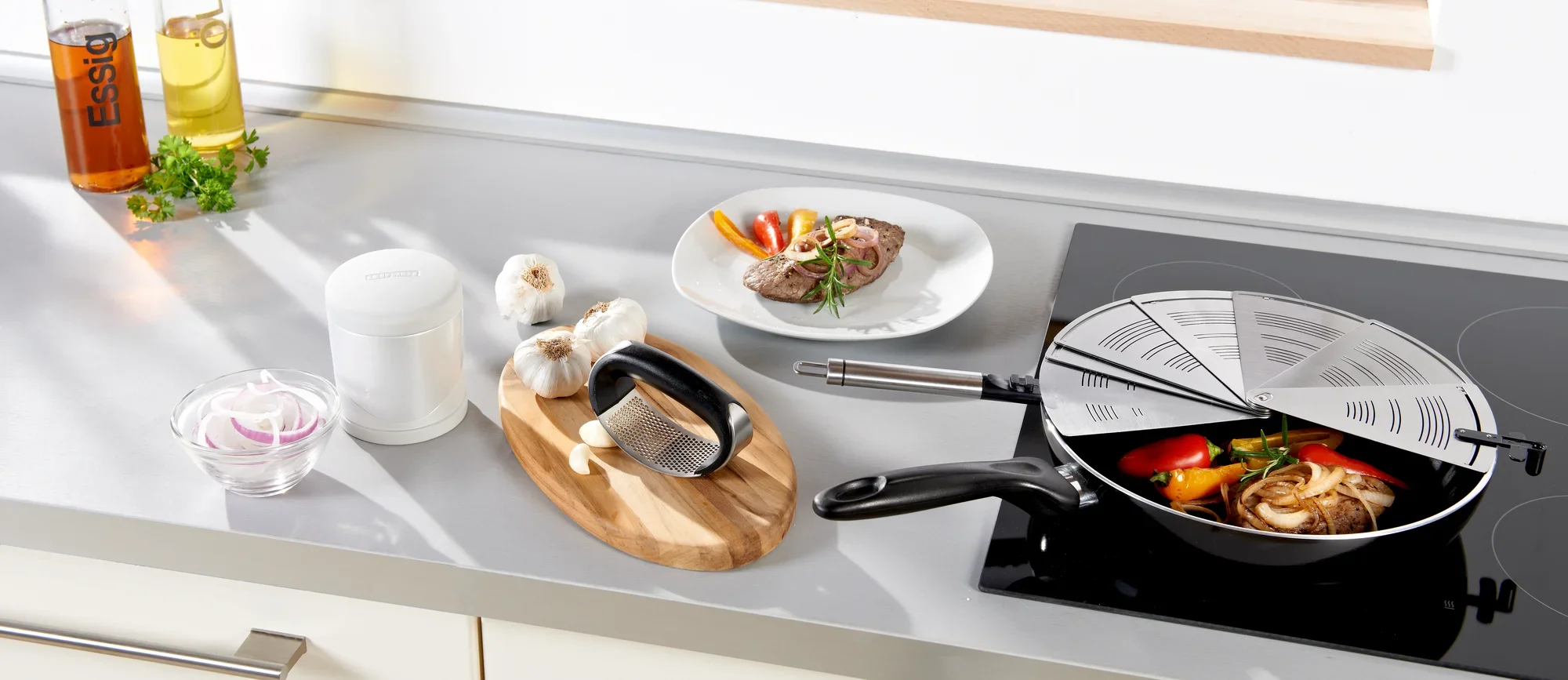 Pare-éclaboussures pliable en acier inoxydable sur une cuisinière, recouvrant une poêle avec des légumes. À côté se trouvent de l'ail, des épices et une assiette avec de la viande. Idéal pour cuisiner proprement.