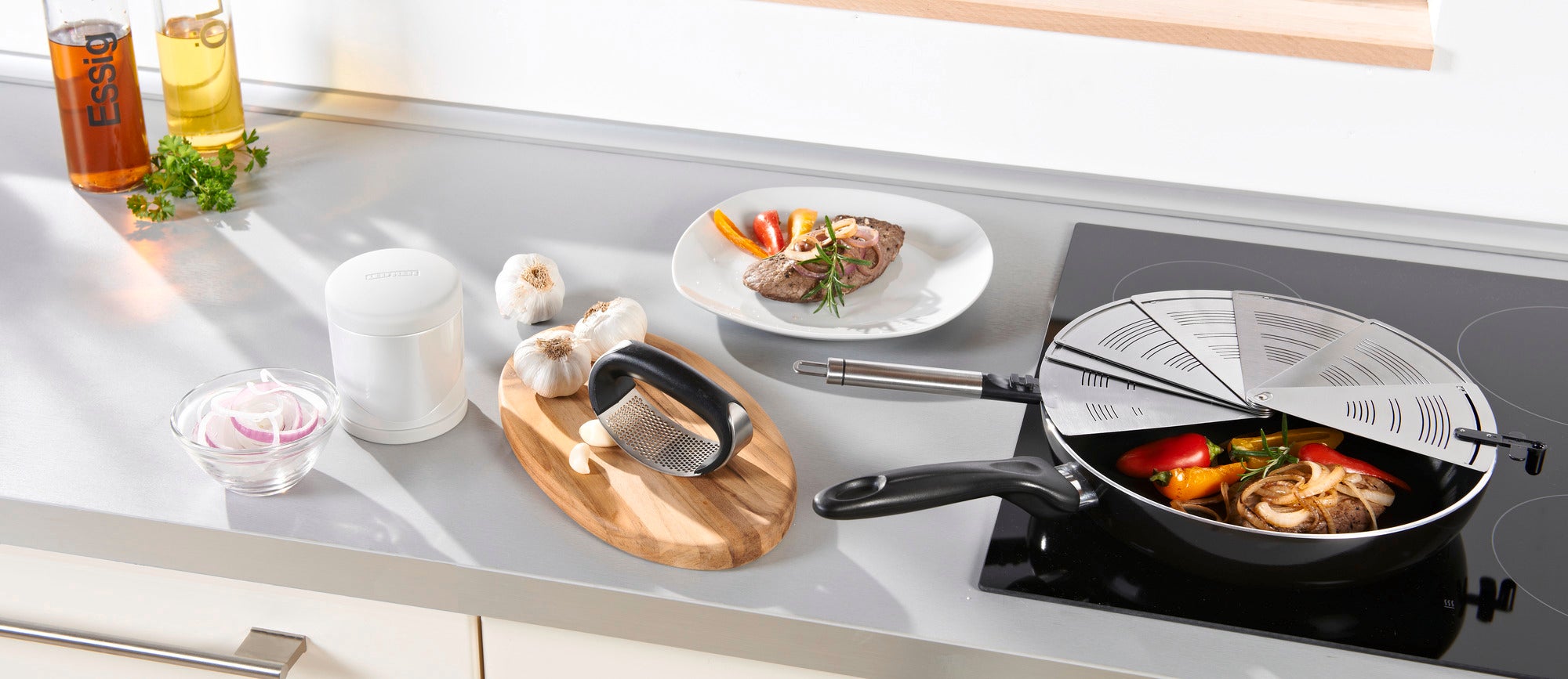 Protector contra salpicaduras plegable de acero inoxidable sobre una cocina, que cubre una sartén con verduras. Junto a ella hay ajo, especias y un plato con carne. Ideal para cocinar sin ensuciar.