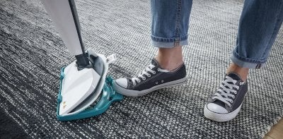 Eine Person in Jeans und Turnschuhen nutzt einen Dampfreiniger, um einen grauen Teppich zu reinigen.