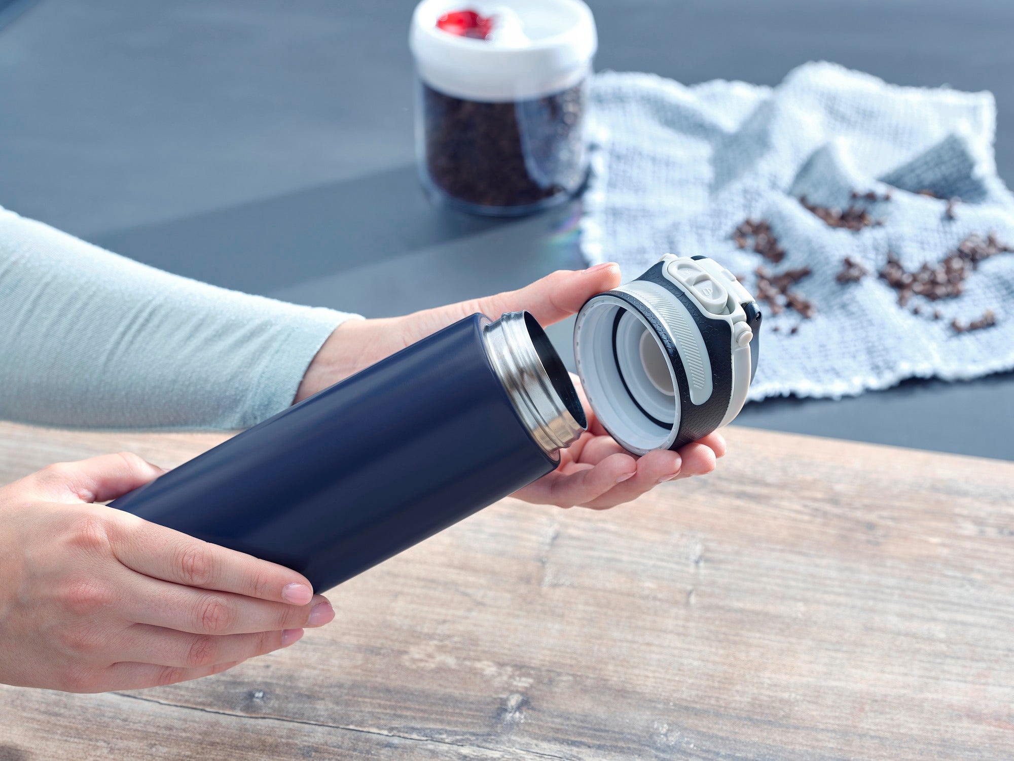 Ein dunkelblauer Isolierbecher Flip mit 600 ml Fassungsvermögen wird geöffnet. Im Hintergrund sind Kaffeebohnen und ein weißer Behälter zu sehen. Ideal für unterwegs.