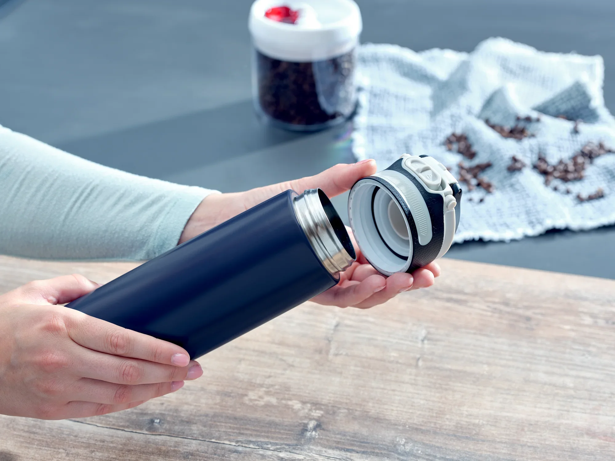 Se abre un vaso térmico Flip azul oscuro con una capacidad de 600 ml. Al fondo se ven granos de café y un recipiente blanco. Ideal para llevar.