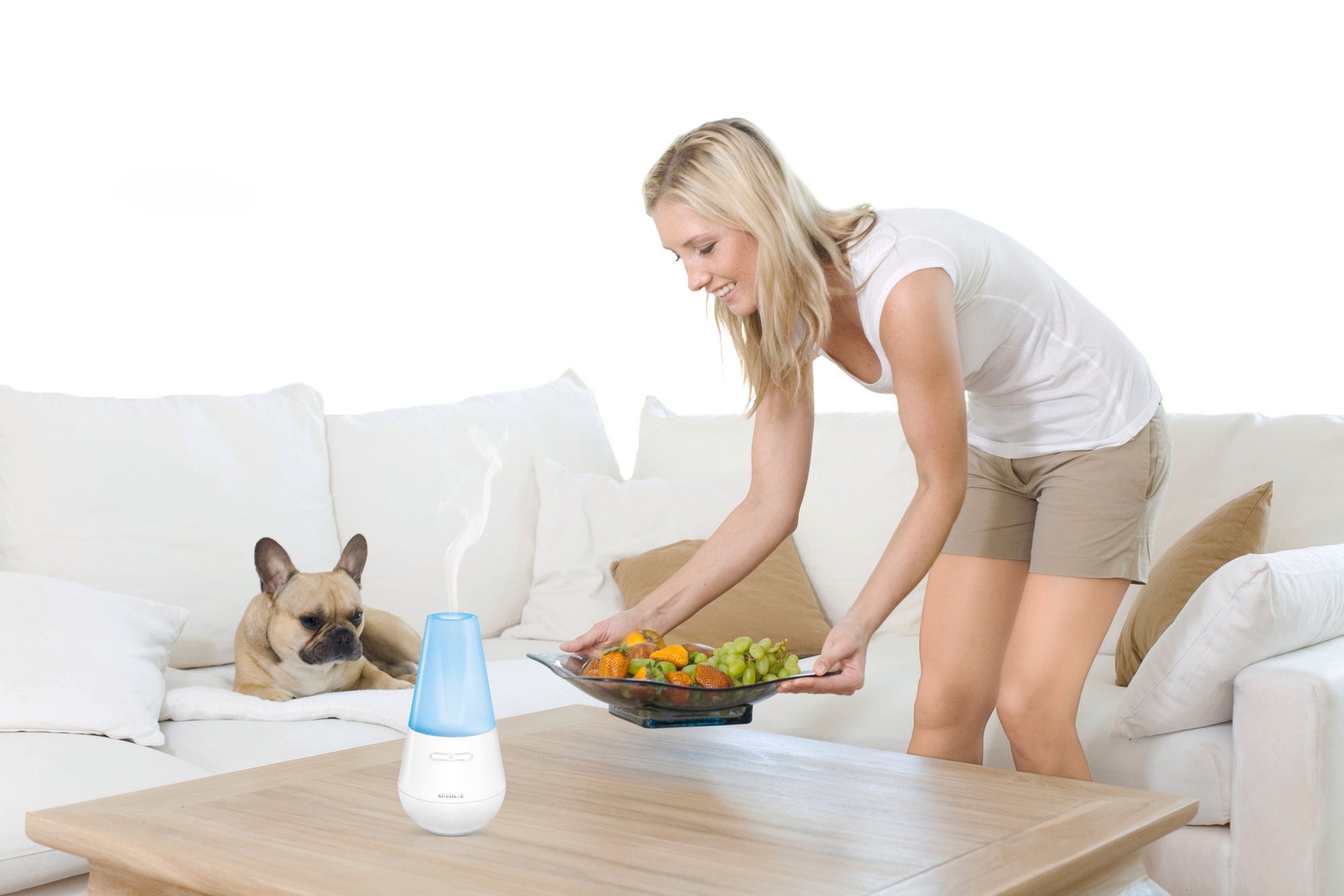 Eine Frau arrangiert eine Obstplatte auf einem Tisch in einem modernen Wohnzimmer. Neben ihr sitzt ein Hund auf dem Sofa. Im Vordergrund steht ein Design Aroma Diffuser Valencia.