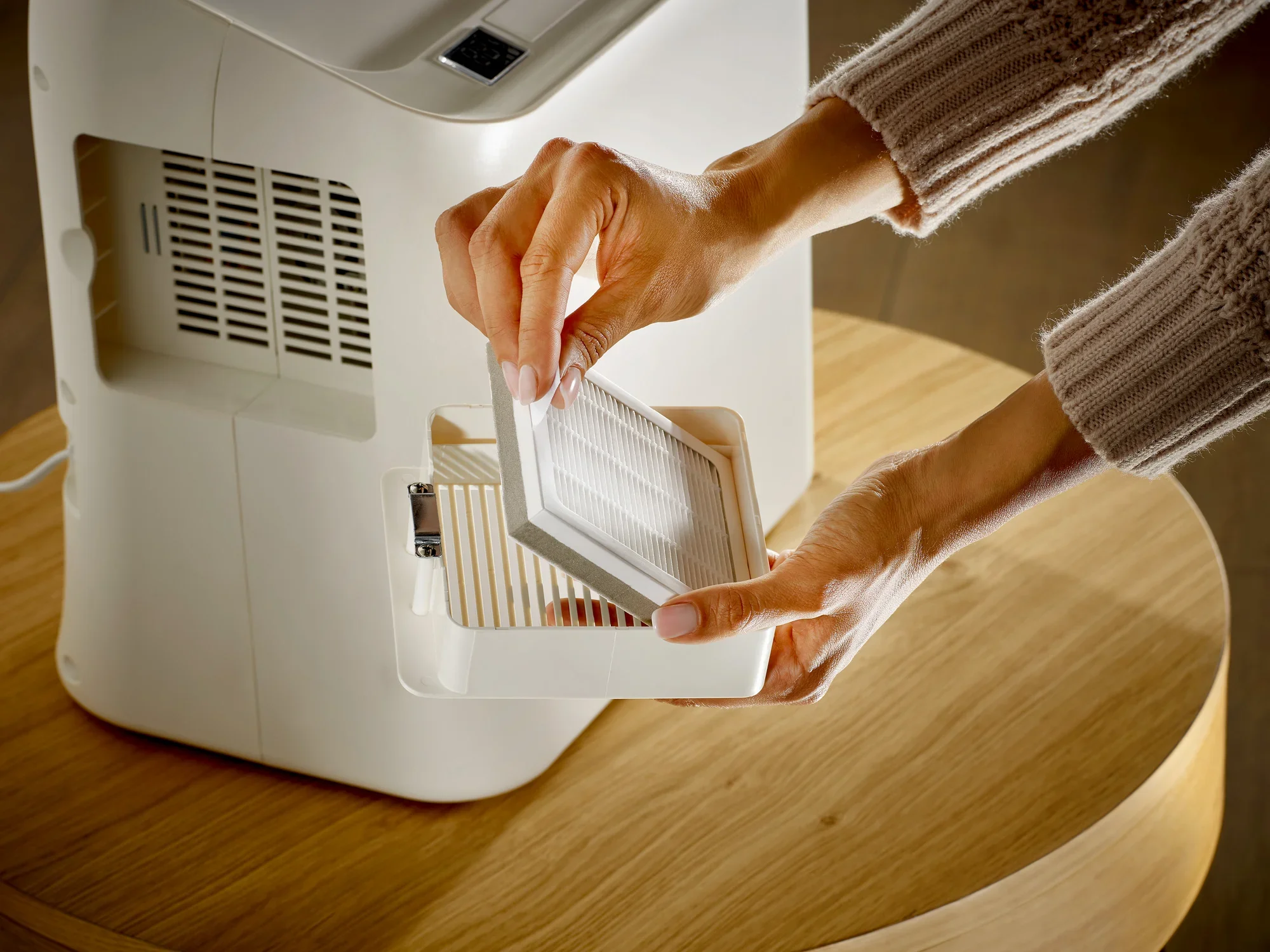 Osoba wymienia filtr oczyszczacza powietrza. Biały oczyszczacz powietrza stoi na drewnianym stole. Ręce pokazują, jak filtr jest używany do poprawy jakości powietrza.