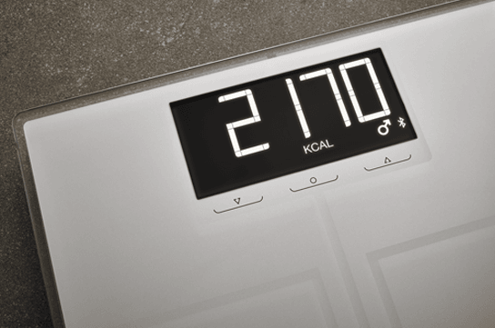 Digitale Waage zeigt 2170 KCAL an, mit Pfeiltasten und Maennlich-Symbol auf dunklem Display vor koernigem Hintergrund.