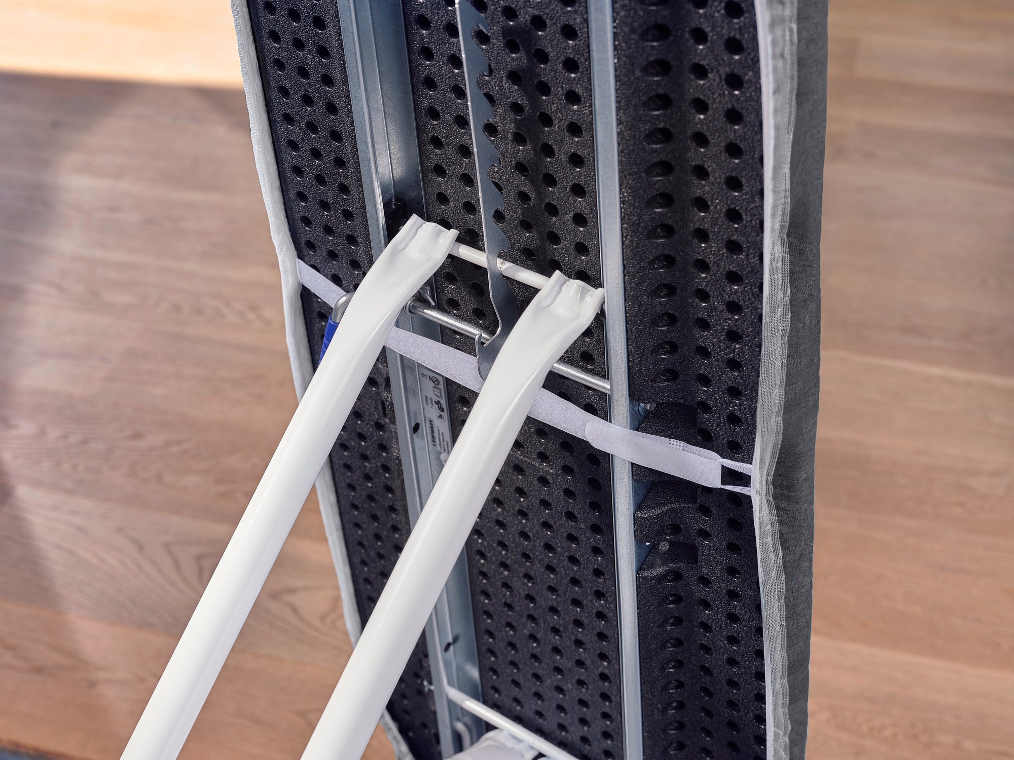 Bügelbrett mit dem Bezug Thermo Reflect Black von unten gezeigt. Der Bezug ist auf einem stabilen Metallrahmen befestigt, der auf einem Holzboden steht.