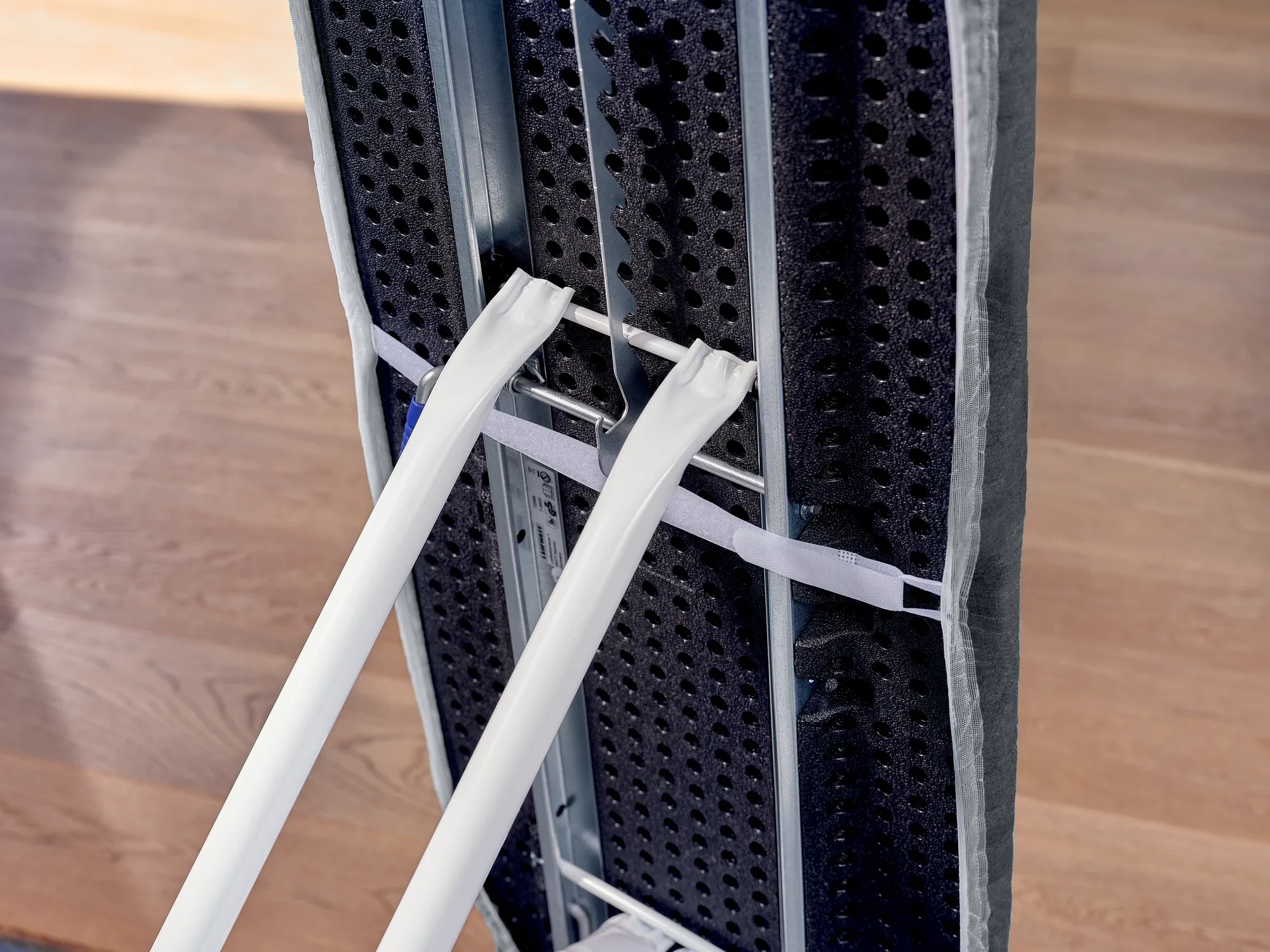 Deska do prasowania z pokrowcem Thermo Reflect Black pokazana od dołu. Pokrowiec jest przymocowany do solidnej metalowej ramy, która stoi na drewnianej podłodze.