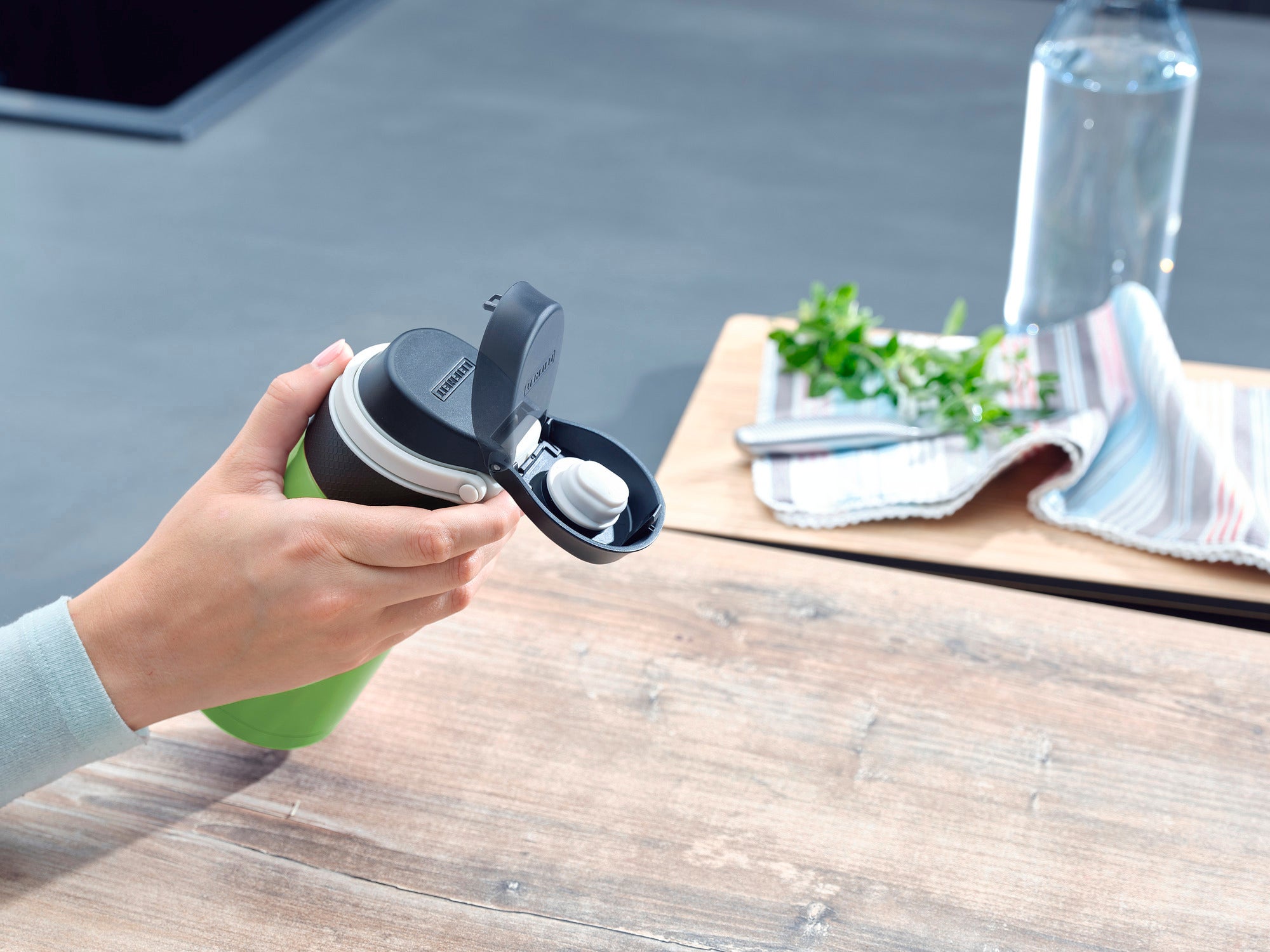 Een persoon houdt een geïsoleerde Flip mok vast in de kleur kiwi met een inhoud van 350 ml. De mok heeft een praktisch klapdeksel. Op de achtergrond zijn een tafel, een theedoek en een waterfles te zien.