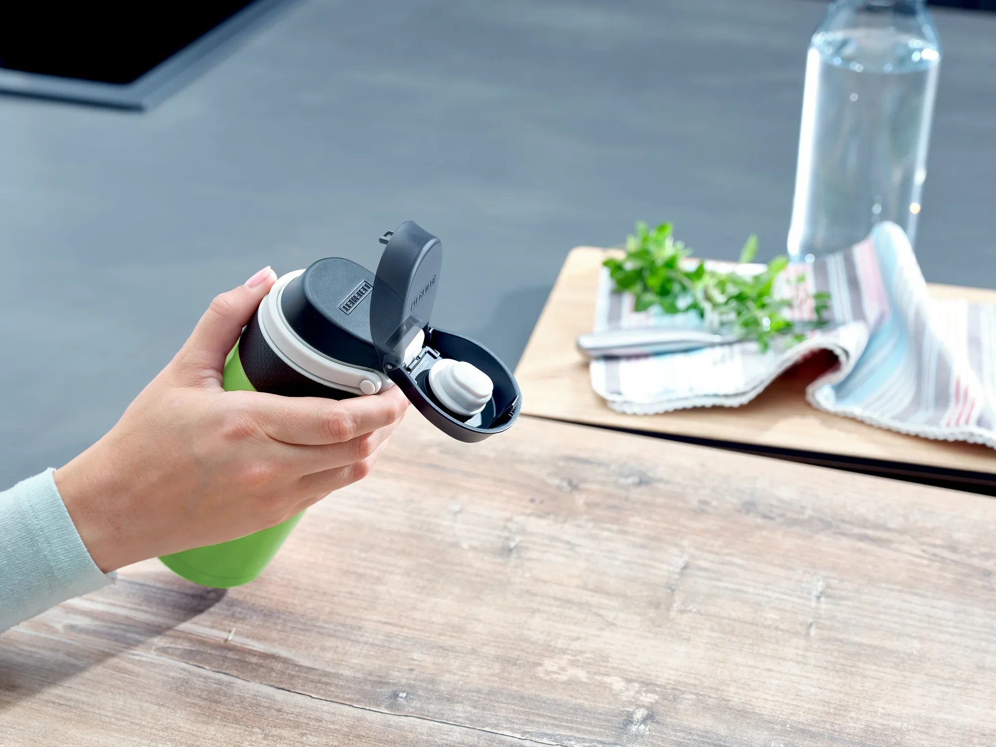 Jedna osoba trzyma izolowany kubek Flip w kolorze kiwi o pojemności 350 ml. Kubek ma praktyczną pokrywkę typu flip-top. W tle widać stół, ściereczkę i butelkę wody.