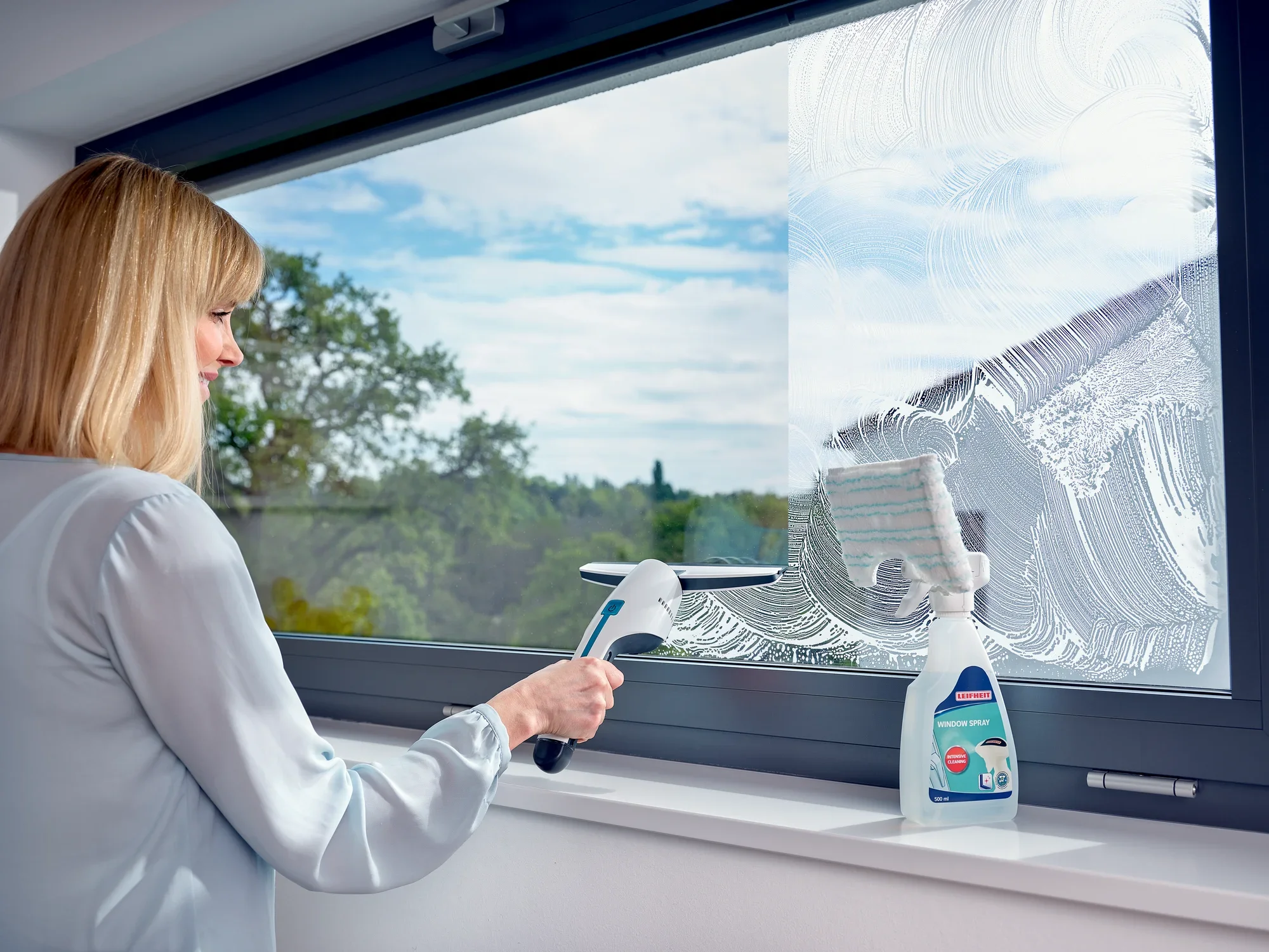 Une femme nettoie une grande fenêtre avec le nettoyeur de vitres Nemo. À côté d'elle se trouve une bouteille de produit nettoyant. La vue vers l'extérieur montre un ciel clair et des arbres.
