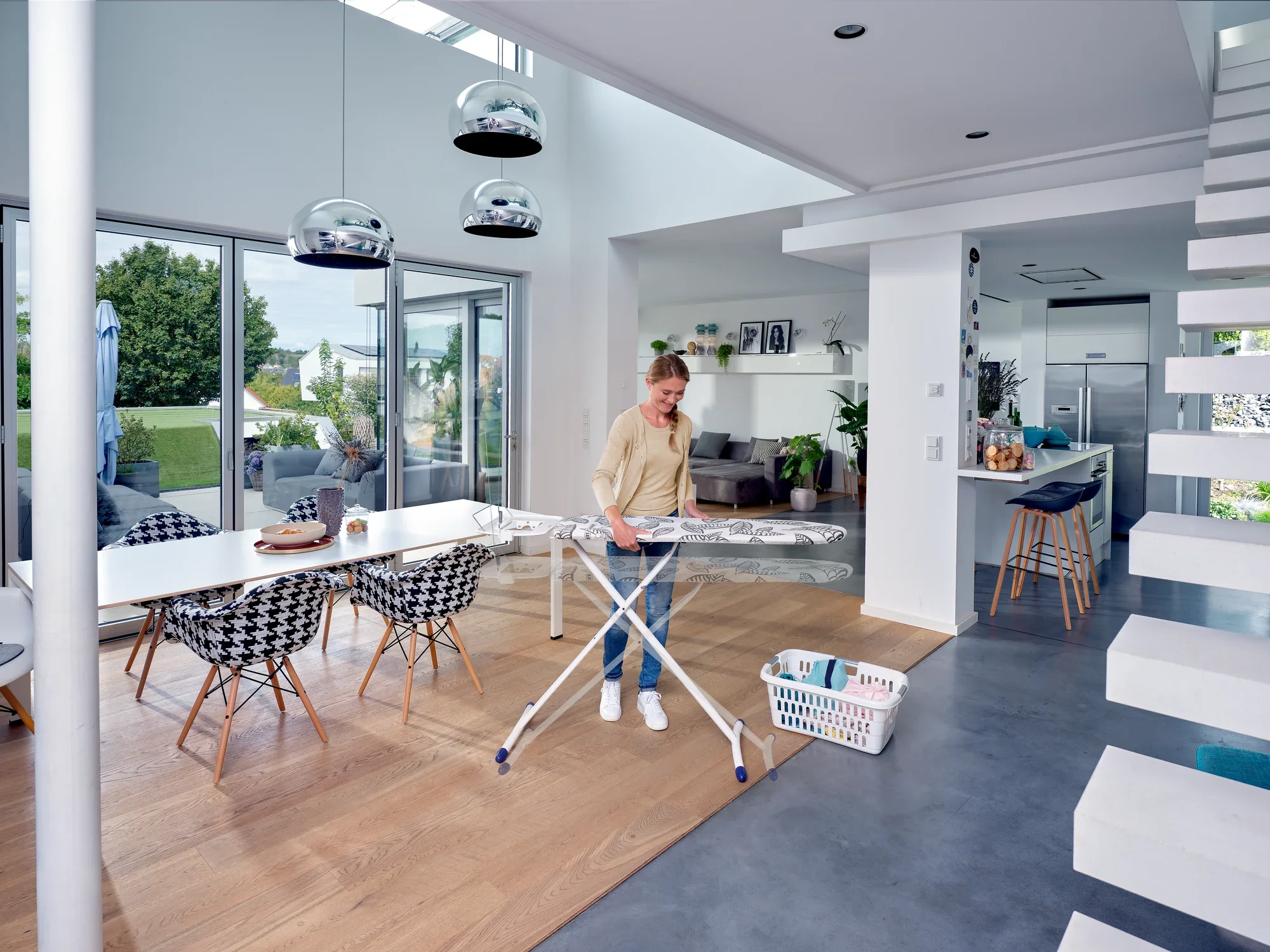Une personne repasse des vêtements sur une table à repasser Air Board Express L Solid MAXX dans un salon moderne et lumineux avec de grandes fenêtres et un aménagement élégant.
