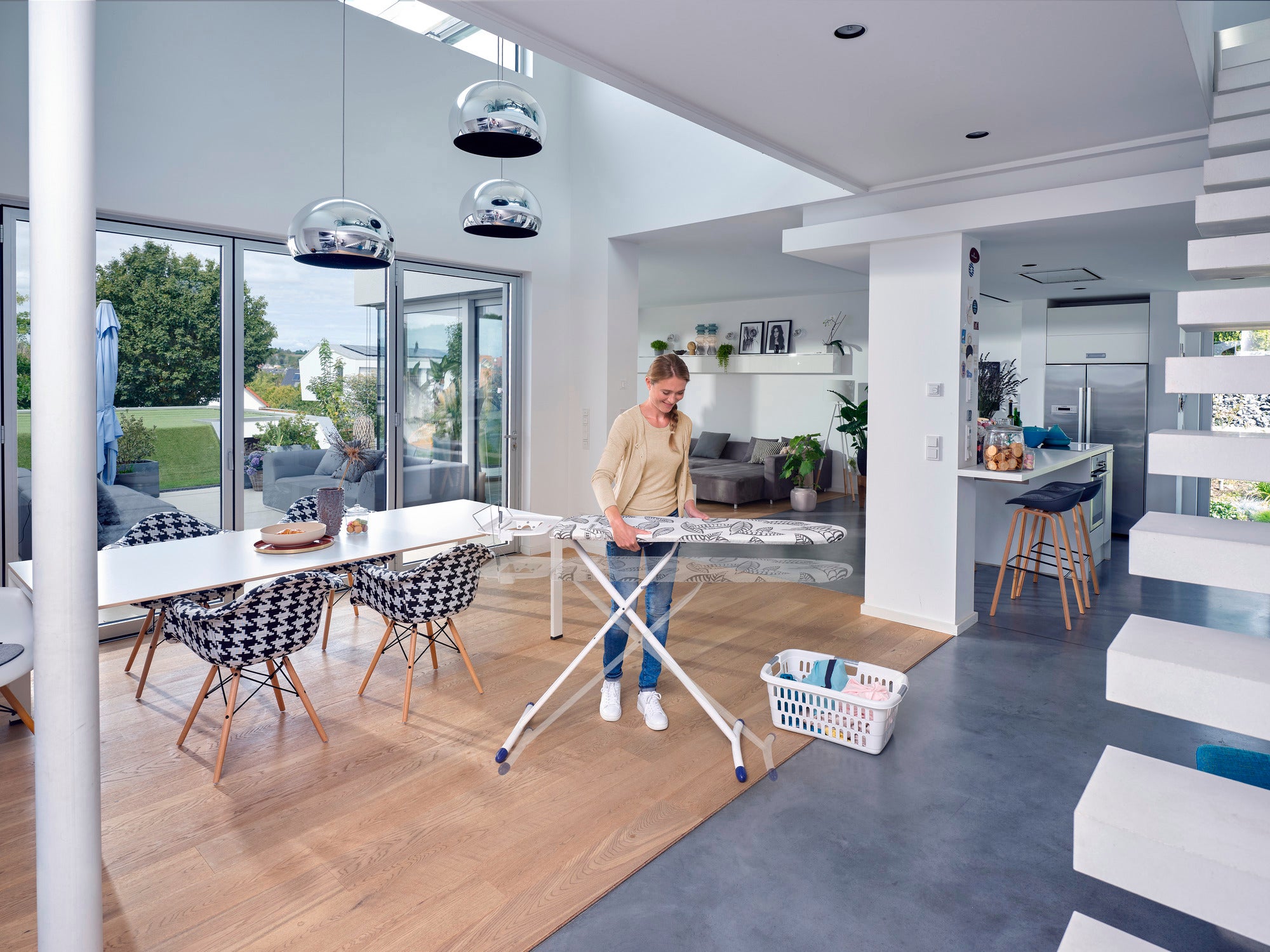 Une personne repasse des vêtements sur une table à repasser Air Board Express L Solid MAXX dans un salon moderne et lumineux avec de grandes fenêtres et un aménagement élégant.