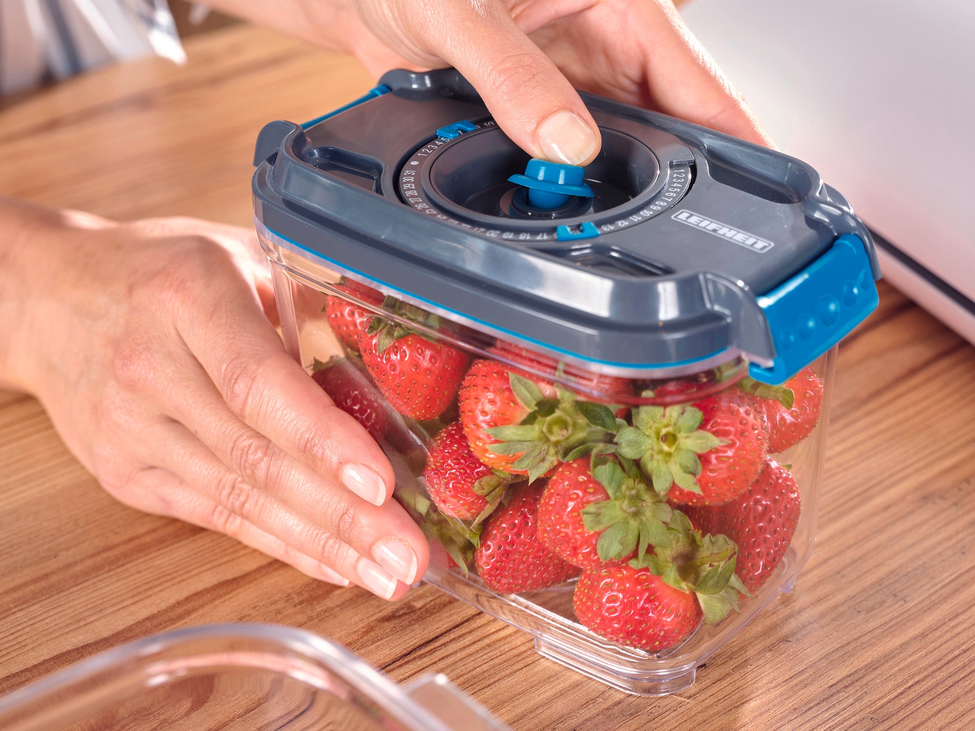 Set vacuüm-voorraadblikken: Een hand houdt een transparant voorraadblik met een blauw deksel, gevuld met verse aardbeien, vast op een houten tafel. Ideaal om voedsel langer te bewaren.