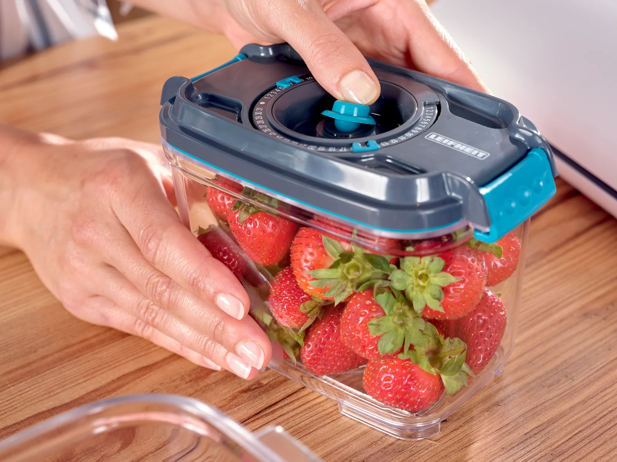 Zestaw próżniowych puszek do przechowywania żywności: Dłoń trzyma na drewnianym stole przezroczystą puszkę do przechowywania żywności z niebieską pokrywką, wypełnioną świeżymi truskawkami. Idealny do dłuższego przechowywania żywności.