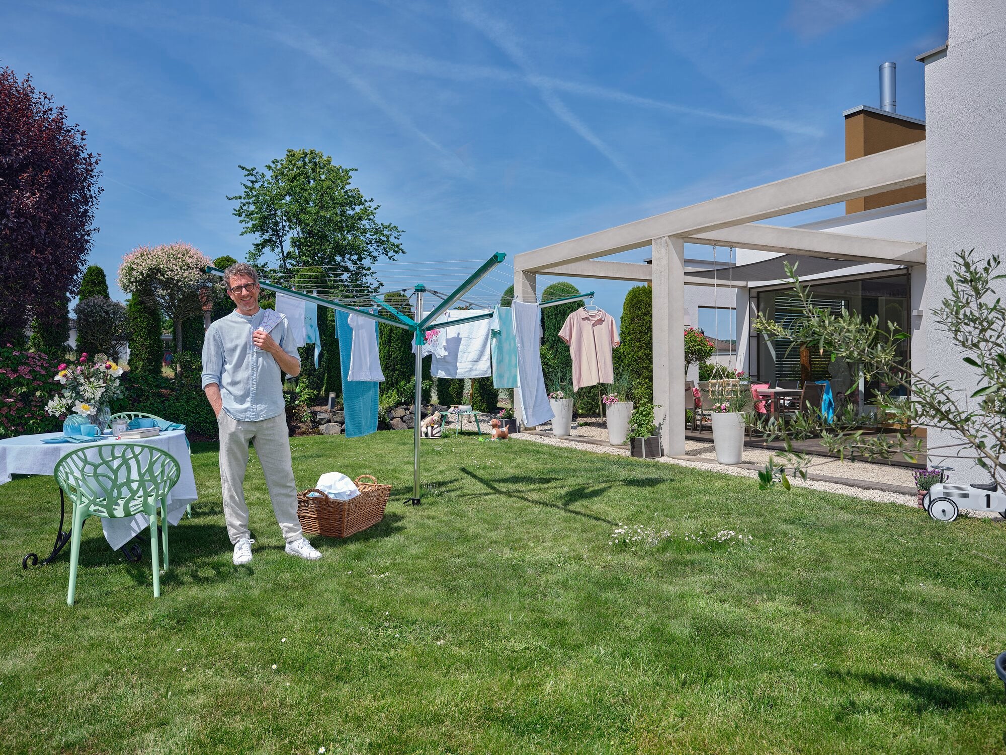 Ein Mann steht in einem gepflegten Garten neben einer Leifheit Linomatic 500 Deluxe Wäschespinne. Die Wäsche hängt zum Trocknen in der Sonne, umgeben von grünen Pflanzen und einem modernen Haus.