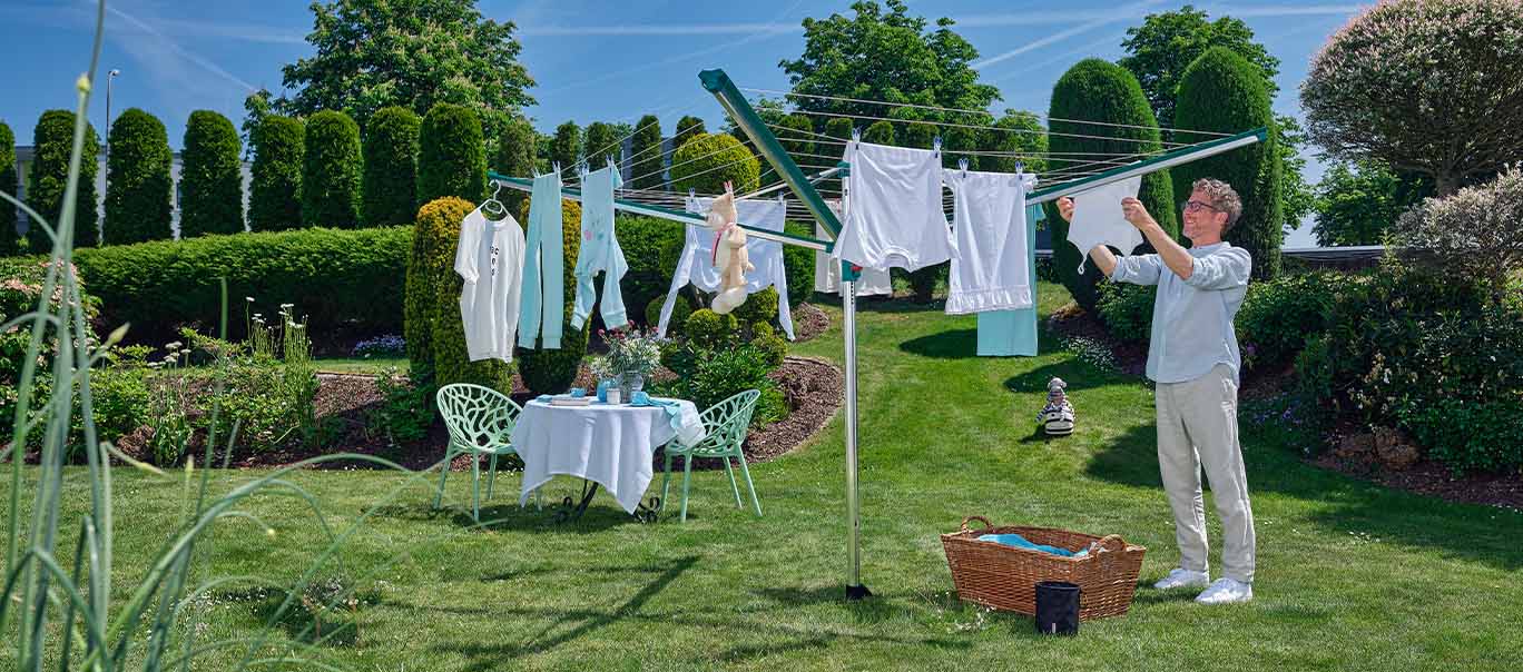 Ein Mann haengt Waesche an einer Leifheit Waeschespinne im sonnigen Garten auf; im Hintergrund ein Tisch mit Stuehlen und bluehenden Pflanzen.