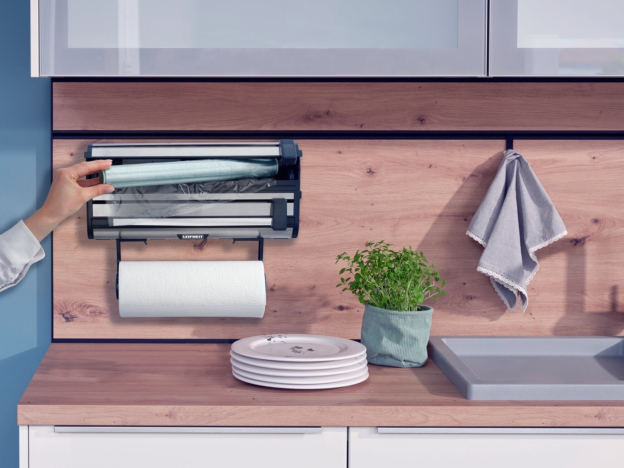 Support mural pour rouleaux Parat Royal monté dans une cuisine moderne. Une main tire un chiffon du support. On aperçoit en dessous des assiettes, un pot d'herbes aromatiques et un torchon.