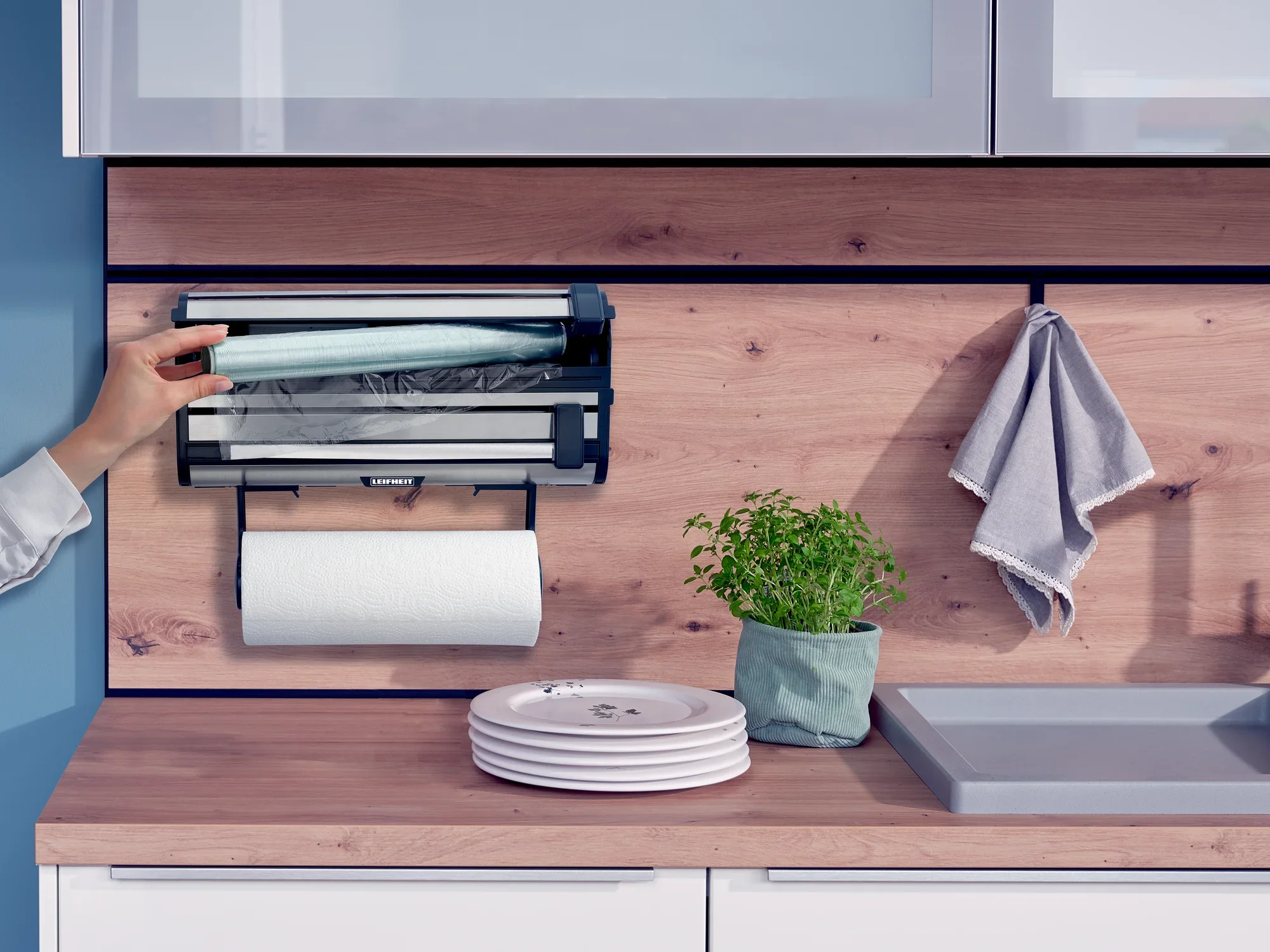 Uchwyt ścienny na rolki Parat Royal zamontowany w nowoczesnej kuchni. Ręka wyciąga ściereczkę z uchwytu. Pod spodem znajdują się talerze, doniczka z ziołami i ręcznik.