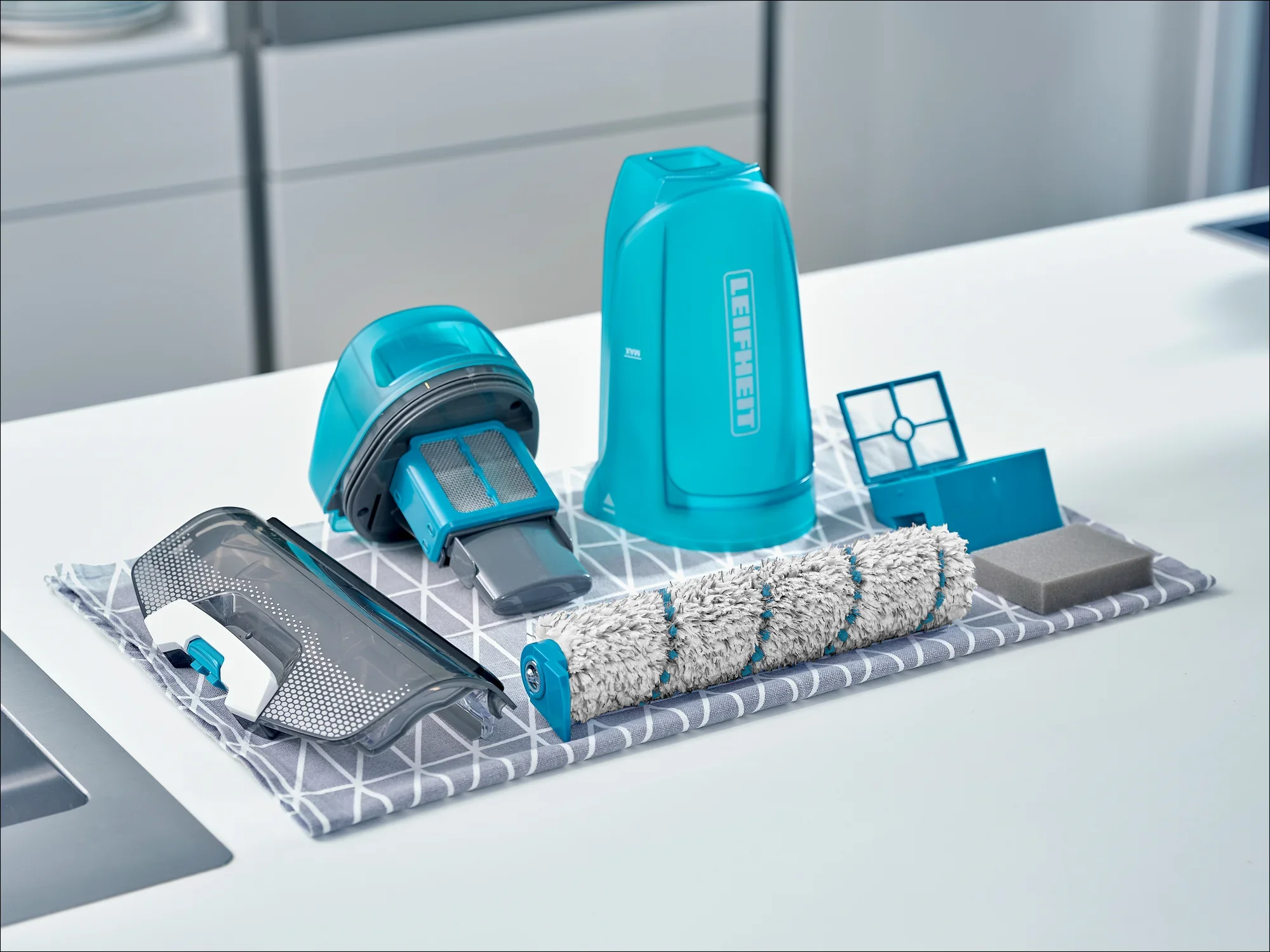 Na stole kuchennym leżą różne części odkurzacza, w tym filtr, rolka szczotki i pojemnik. Części są koloru niebieskiego i białego.