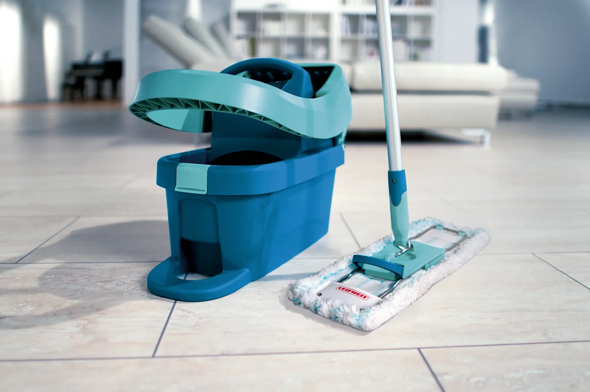 A turquoise Profi XL mop with matching bucket on a light-coloured tiled floor. A modern living room with a white sofa can be seen in the background.