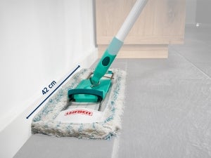 Een vloerwisser met een turquoise handvat en een witte, pluizige doek reinigt een hoekje van een grijze tegelvloer.