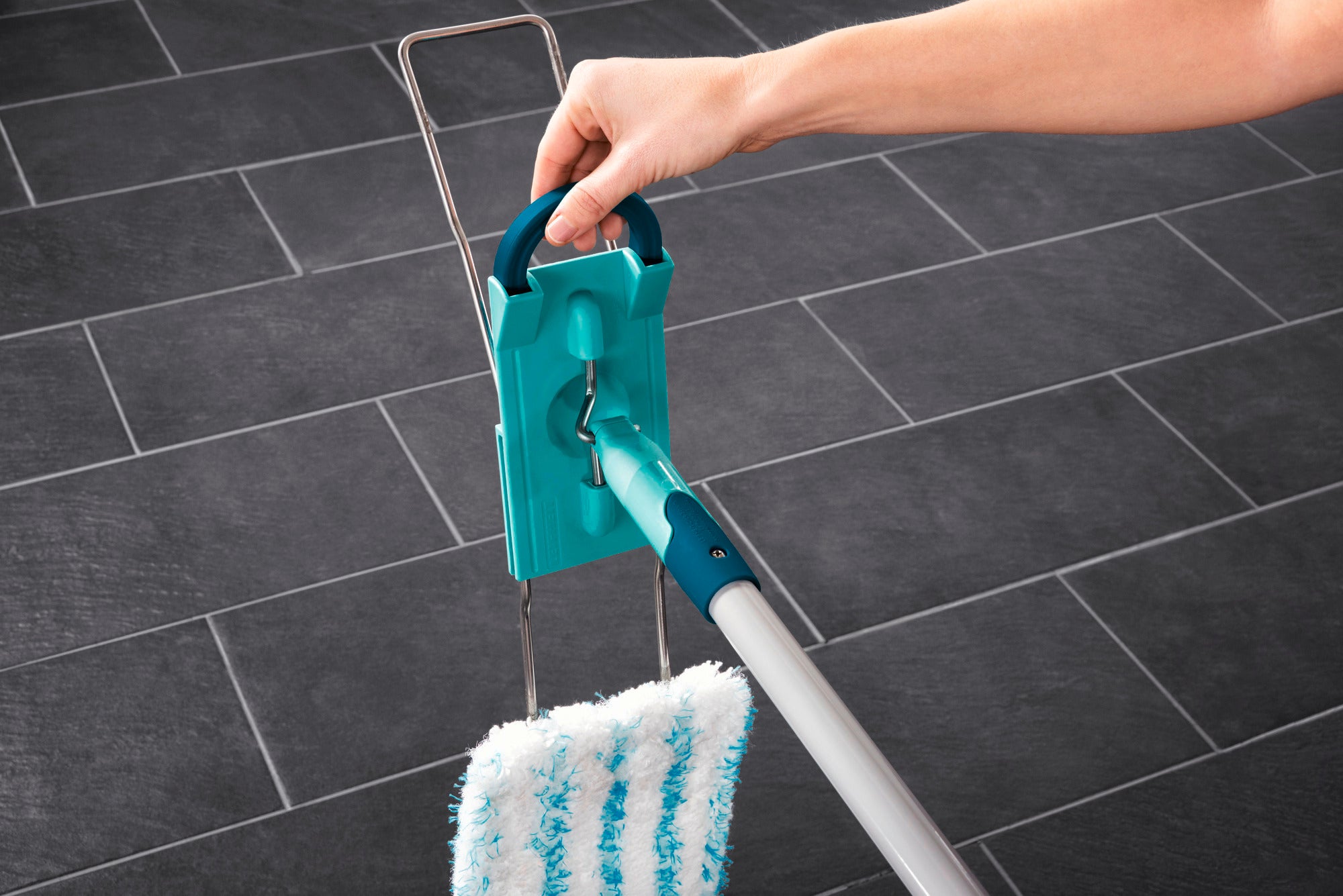 Un balai Profi XL avec un manche en acier est utilisé sur du carrelage foncé. Une main tient le manche du balai, qui est équipé d'une tête de nettoyage verte.