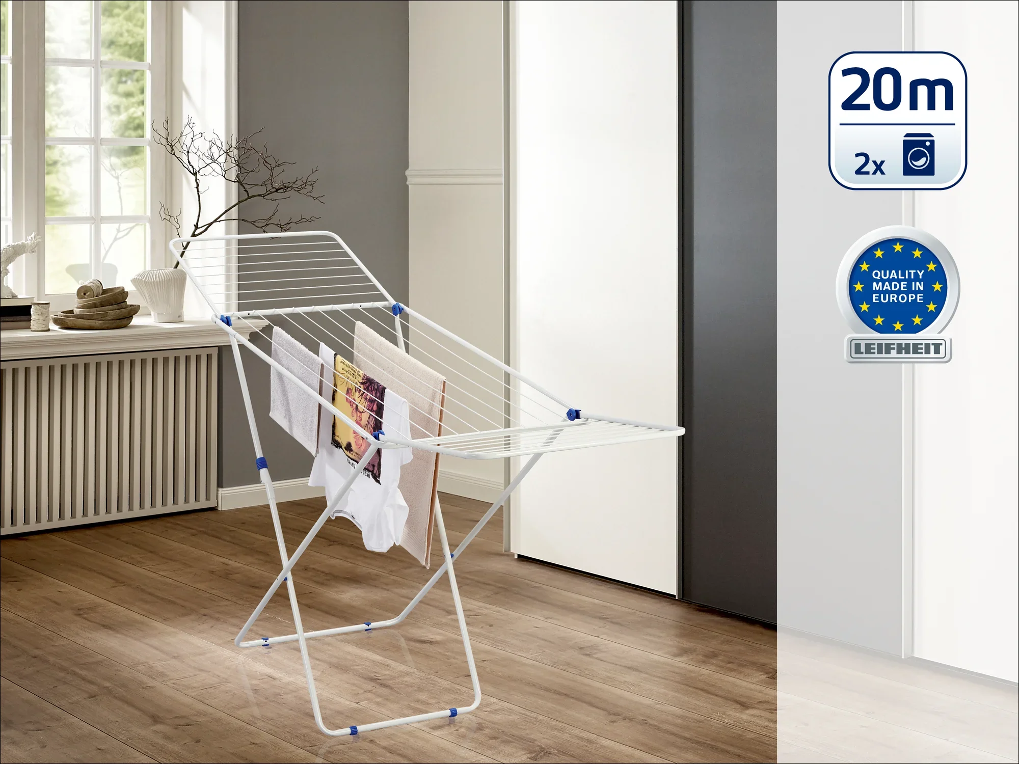 Biała suszarka stojąca na podłodze stoi w nowoczesnym pomieszczeniu z drewnianą podłogą i dużymi oknami. Oferuje 20 metrów długości suszenia i została wyróżniona znakiem "Quality Made in Europe".