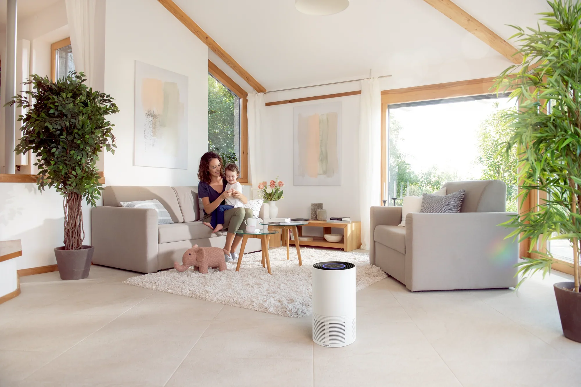 Kobieta siedzi na sofie z dzieckiem w jasnym salonie. Na pierwszym planie znajduje się oczyszczacz powietrza Airfresh Clean 400, który zapewnia czyste powietrze.