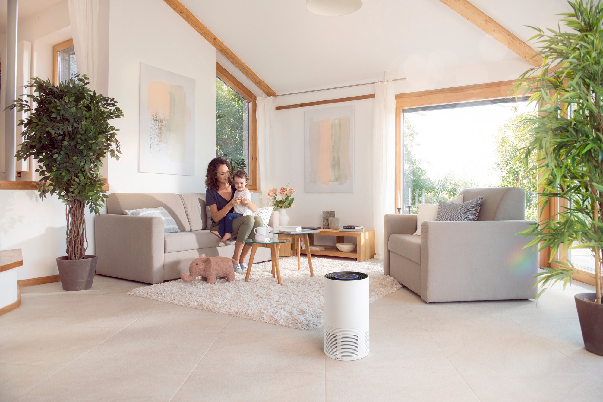 A woman sits with a child on a sofa in a bright living room. In the foreground is the Airfresh Clean 400 air purifier, which ensures clean air.