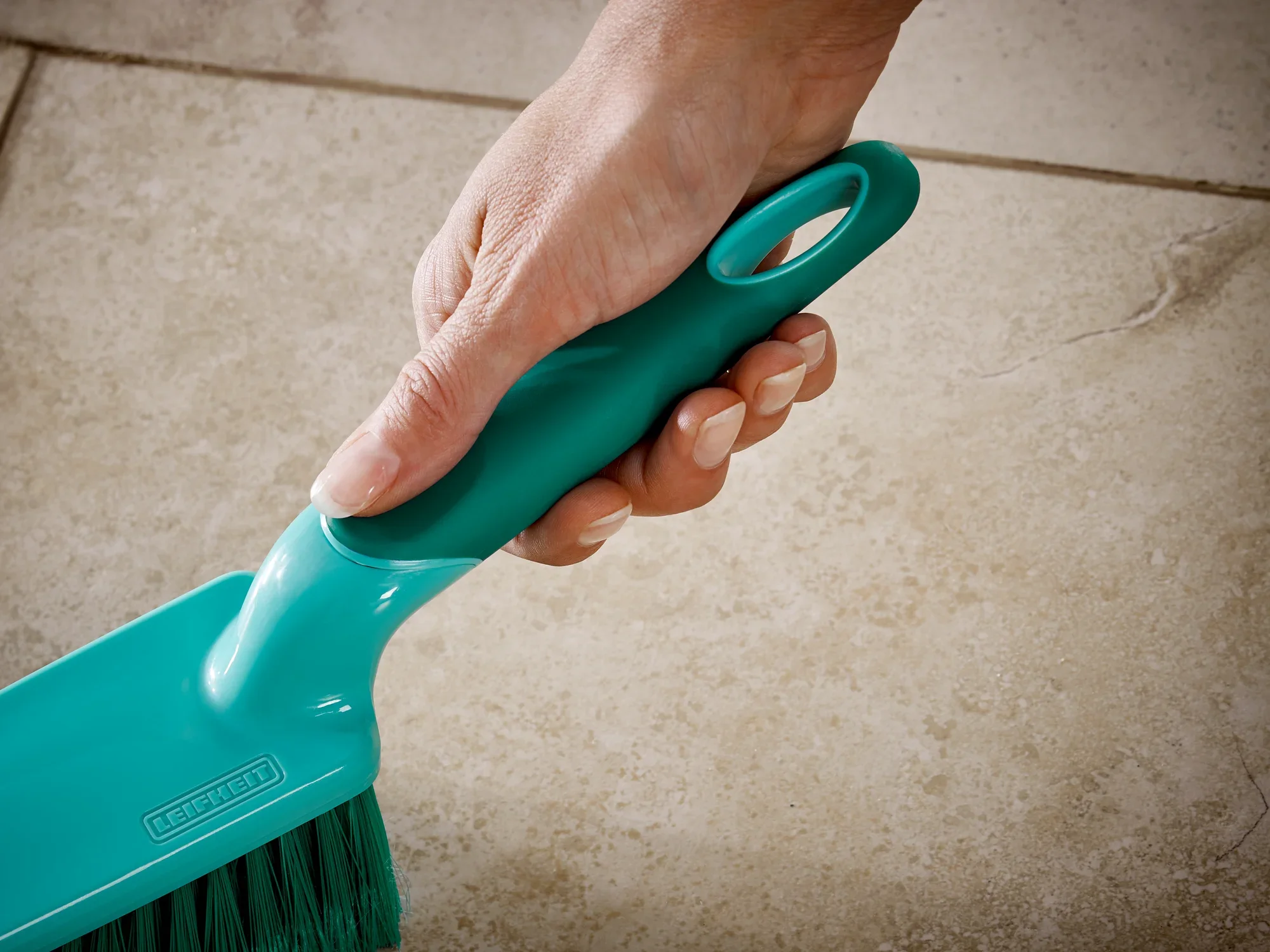 Eine Hand hält ein Leifheit Handkehrset mit einem blauen Griff und Borsten, ideal für die Reinigung von Fliesenböden. Das ergonomische Design erleichtert die Handhabung.