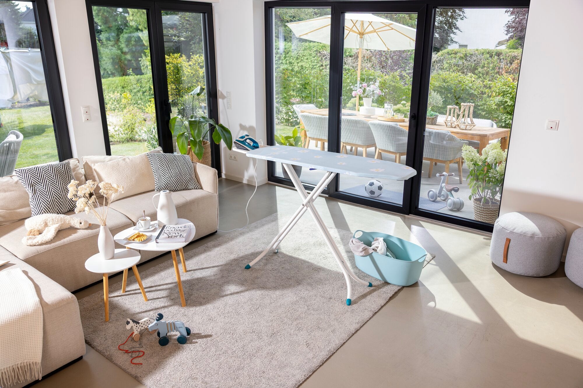 Bügelbrett Leifheit Classic M Compact in einem modernen Wohnzimmer mit großen Fenstern. Das Brett steht auf einem Teppich, umgeben von Möbeln und Dekorationen.
