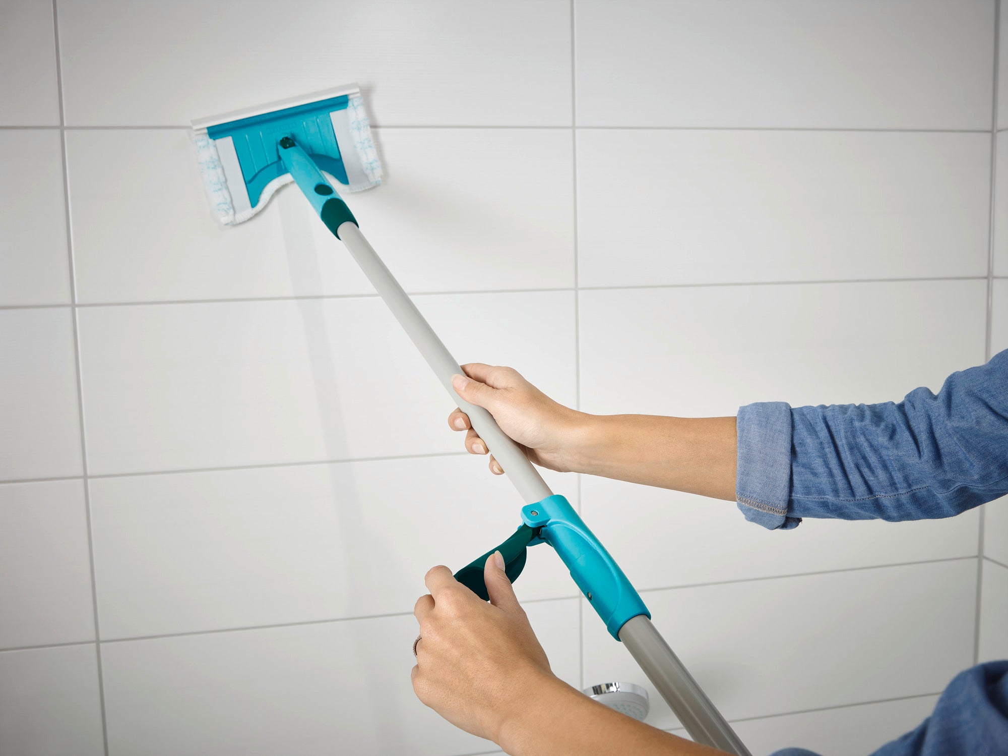 A Flexipad tile and tub squeegee with a telescopic handle is being used by a person to clean white tiles. The squeegee has an adjustable handle for hard-to-reach areas.