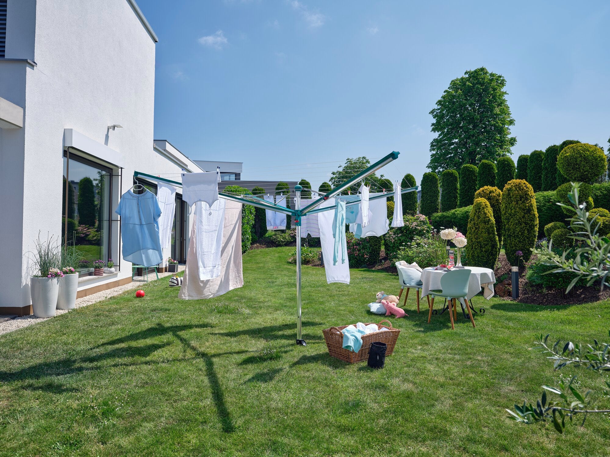 Ein Wäscheschirm Linomatic 600 Deluxe steht im Garten eines modernen Hauses. Frische Wäsche hängt zum Trocknen, während ein Tisch mit Getränken und ein Korb daneben stehen.