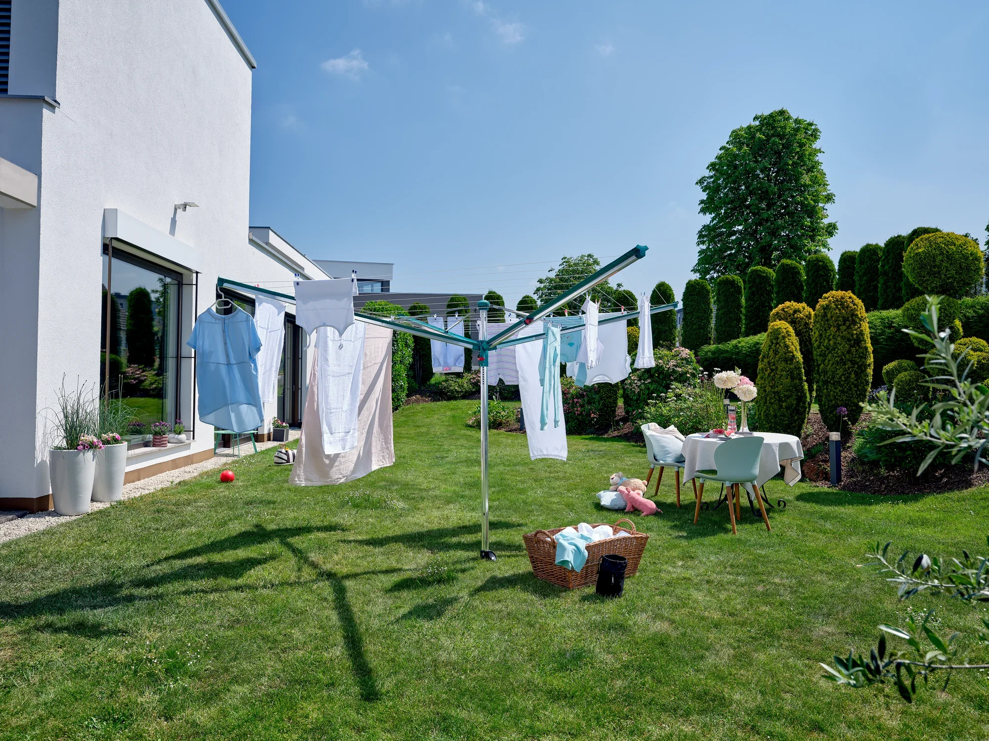 Parasol na pranie Linomatic 600 Deluxe stoi w ogrodzie nowoczesnego domu. Świeże pranie wisi do wyschnięcia, a obok stoi stolik z napojami i kosz.