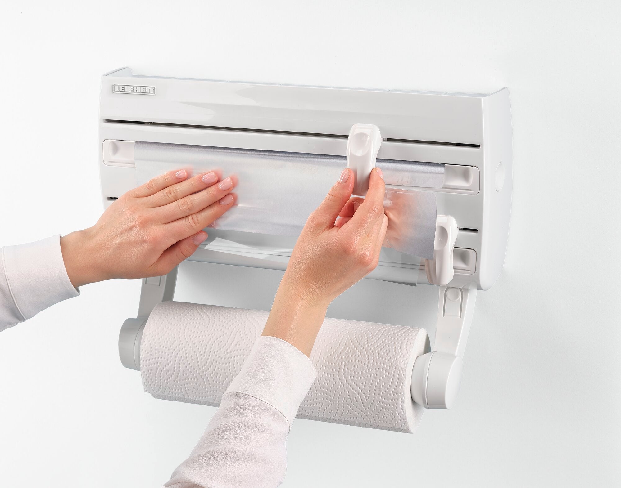 A Parat F2 wall-mounted roll holder from Leifheit is mounted on a white wall. Two hands pull up the transparent cover to remove the kitchen roll.