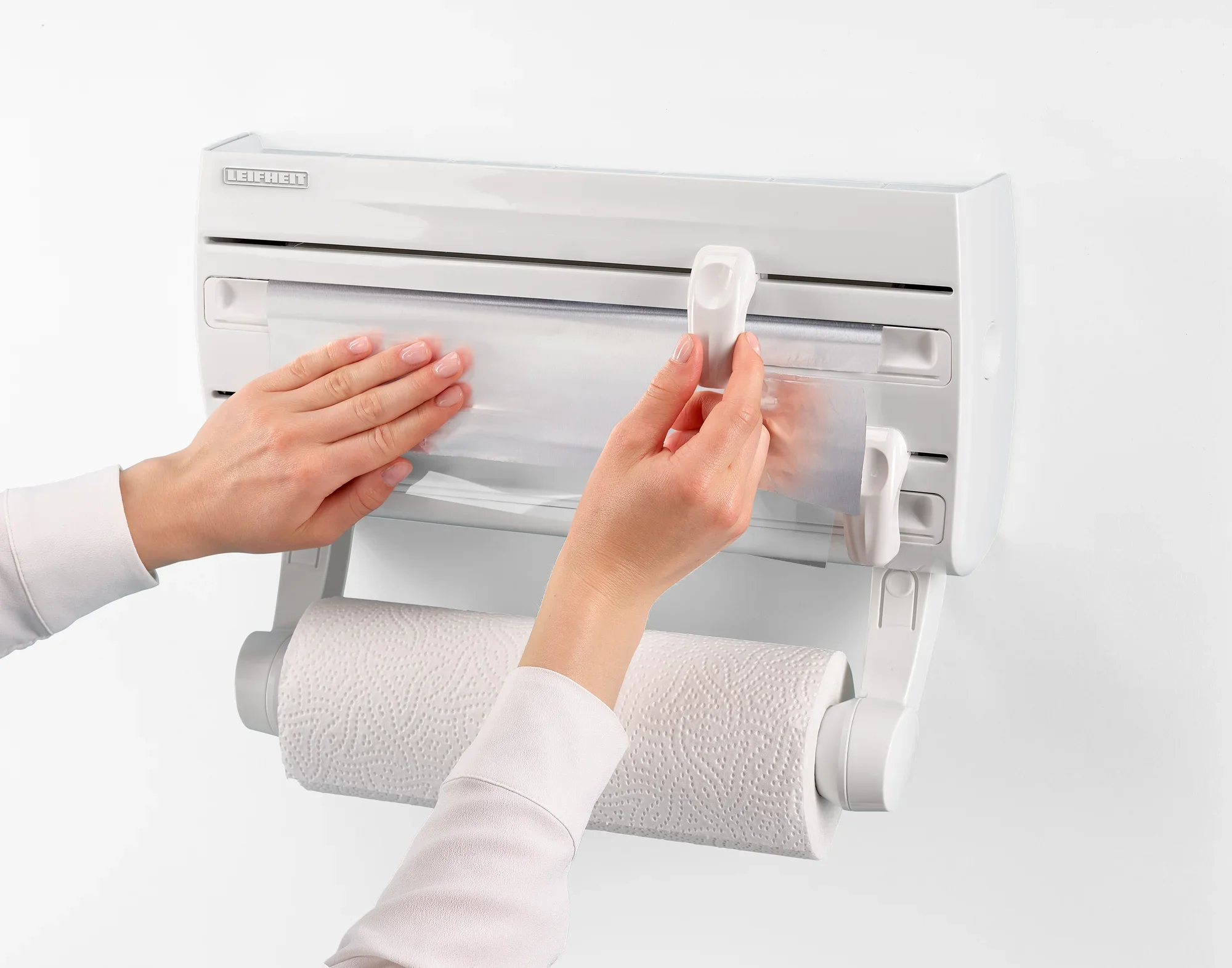 Un portarotoli da parete Parat F2 di Leifheit è montato su una parete bianca. Due mani sollevano il coperchio trasparente per estrarre il rotolo da cucina.