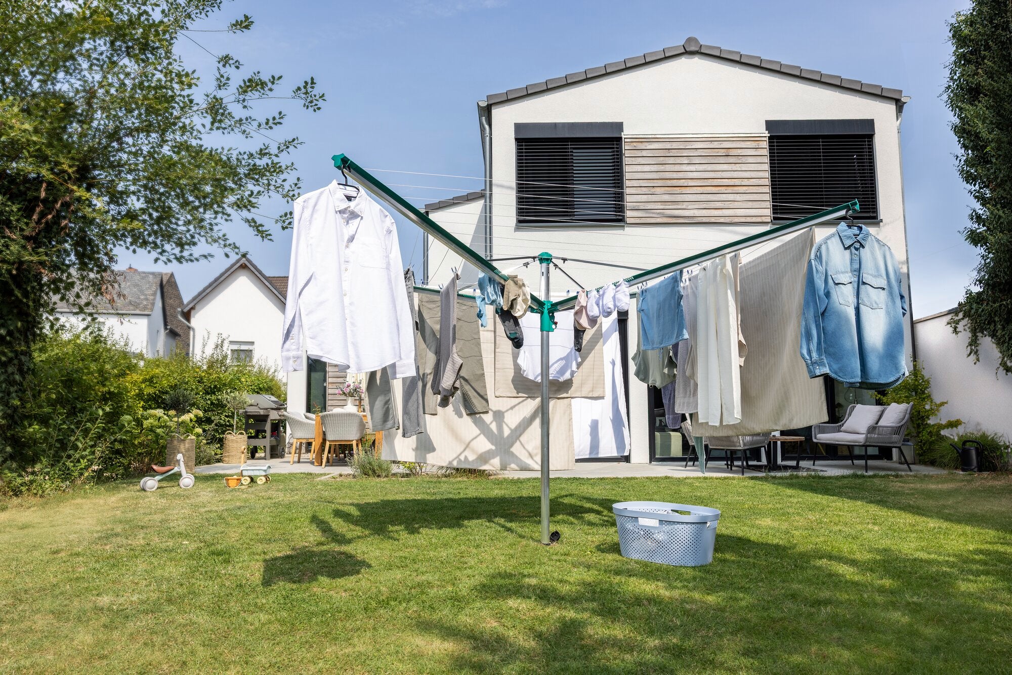 Wäschespinne Linomatic 600 Deluxe von Leifheit im Einsatz in einem sonnigen Garten. Verschiedene Kleidungsstücke hängen zum Trocknen an der Wäschespinne. Im Hintergrund ist ein modernes Haus zu sehen.
