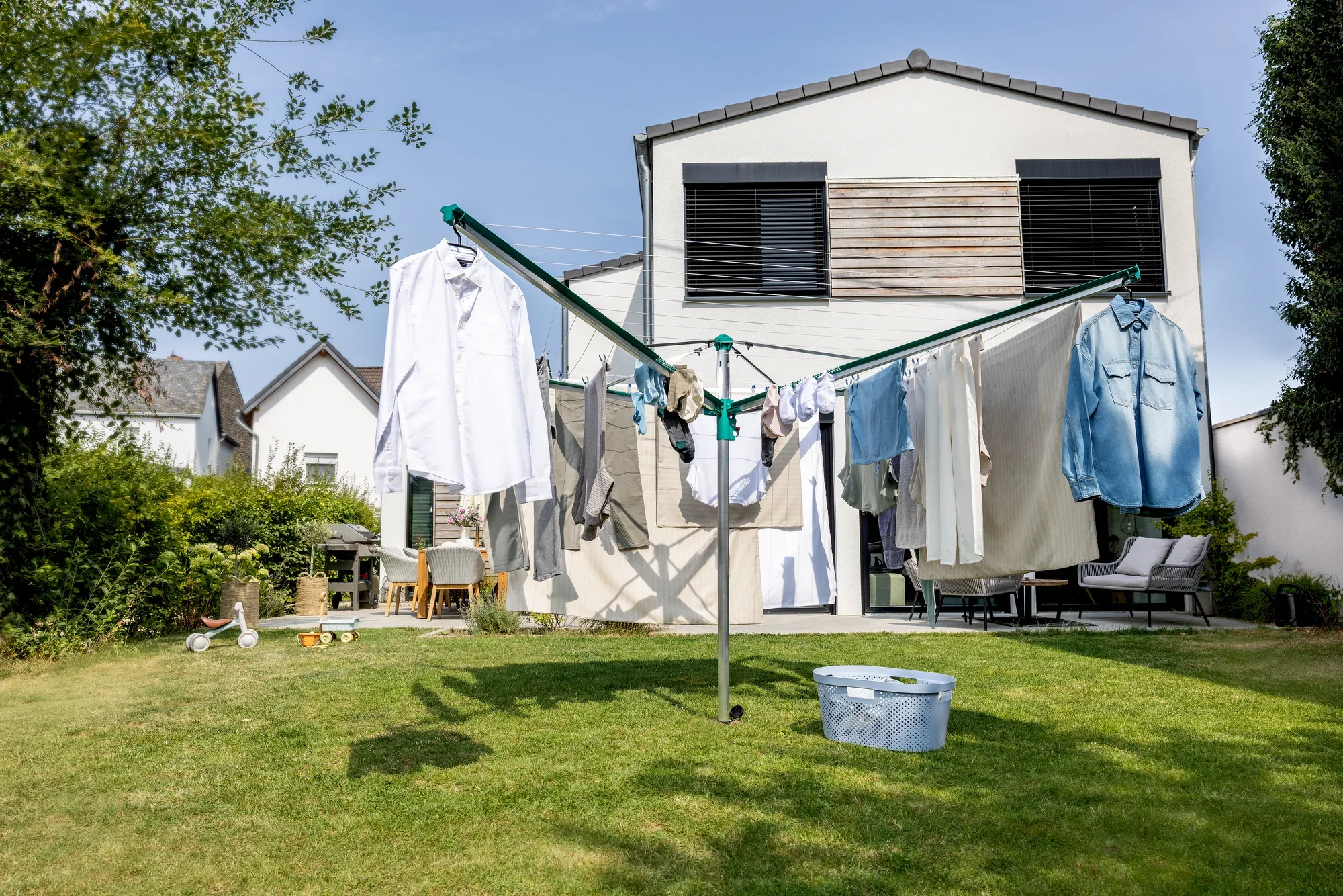 Wäschespinne Linomatic 600 Deluxe von Leifheit im Einsatz in einem sonnigen Garten. Verschiedene Kleidungsstücke hängen zum Trocknen an der Wäschespinne. Im Hintergrund ist ein modernes Haus zu sehen.