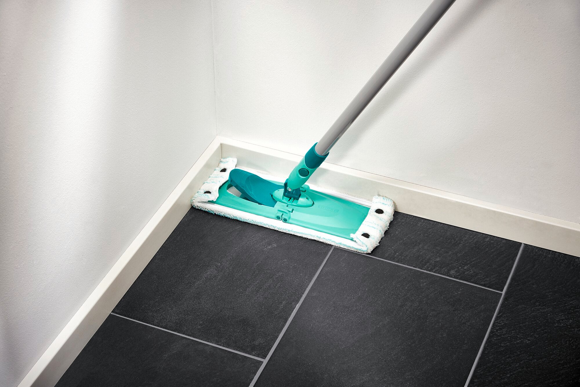 Ein Bodenwischer der Marke Combi M reinigt eine Ecke eines Raumes mit dunklen Fliesen. Der Wischer hat einen türkisfarbenen Kopf und einen grauen Stiel, ideal für gründliche Reinigung.