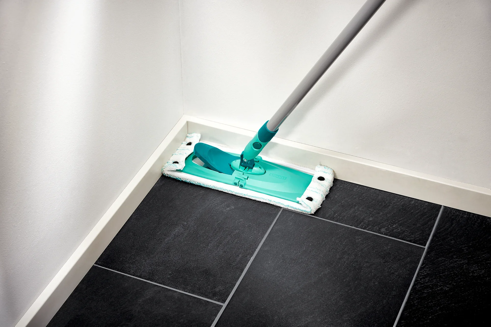 A floor mop from the Combi M series cleans a corner of a room with dark tiles. The mop has a turquoise head and a grey handle, ideal for thorough cleaning.