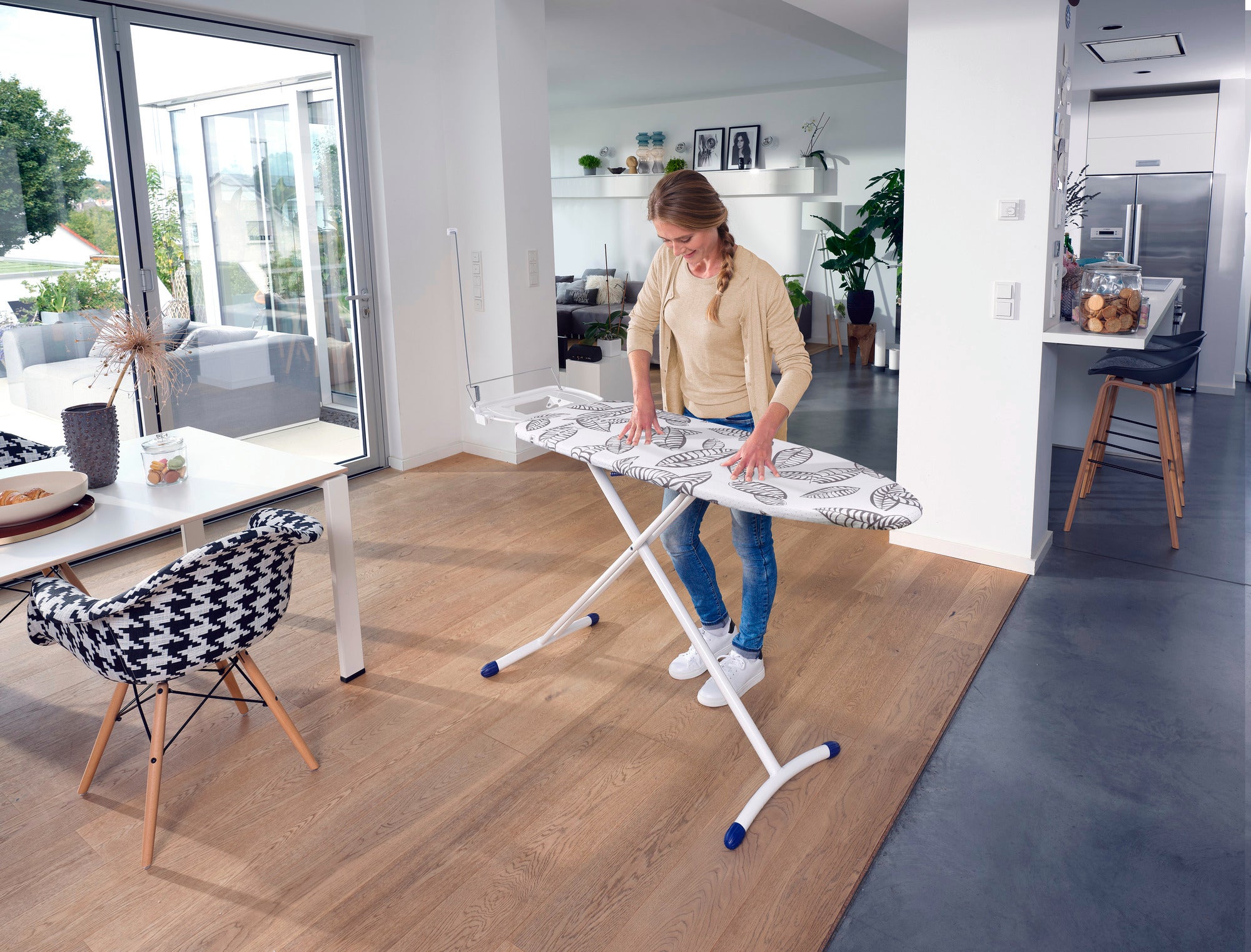 Eine Frau steht in einem modernen, hellen Wohnzimmer und nutzt das Bügelbrett Air Board Express L Solid MAXX. Das Brett hat ein elegantes Design und bietet eine große Bügelfläche.