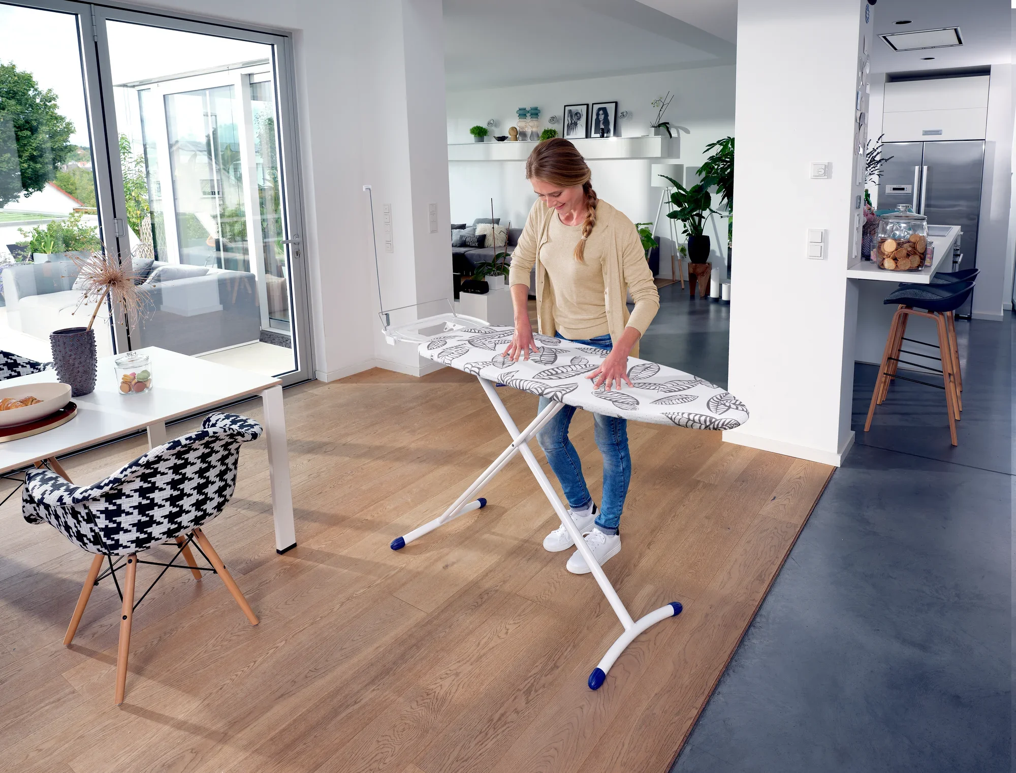 Une femme se tient dans un salon moderne et lumineux et utilise la table à repasser Air Board Express L Solid MAXX. La table a un design élégant et offre une grande surface de repassage.