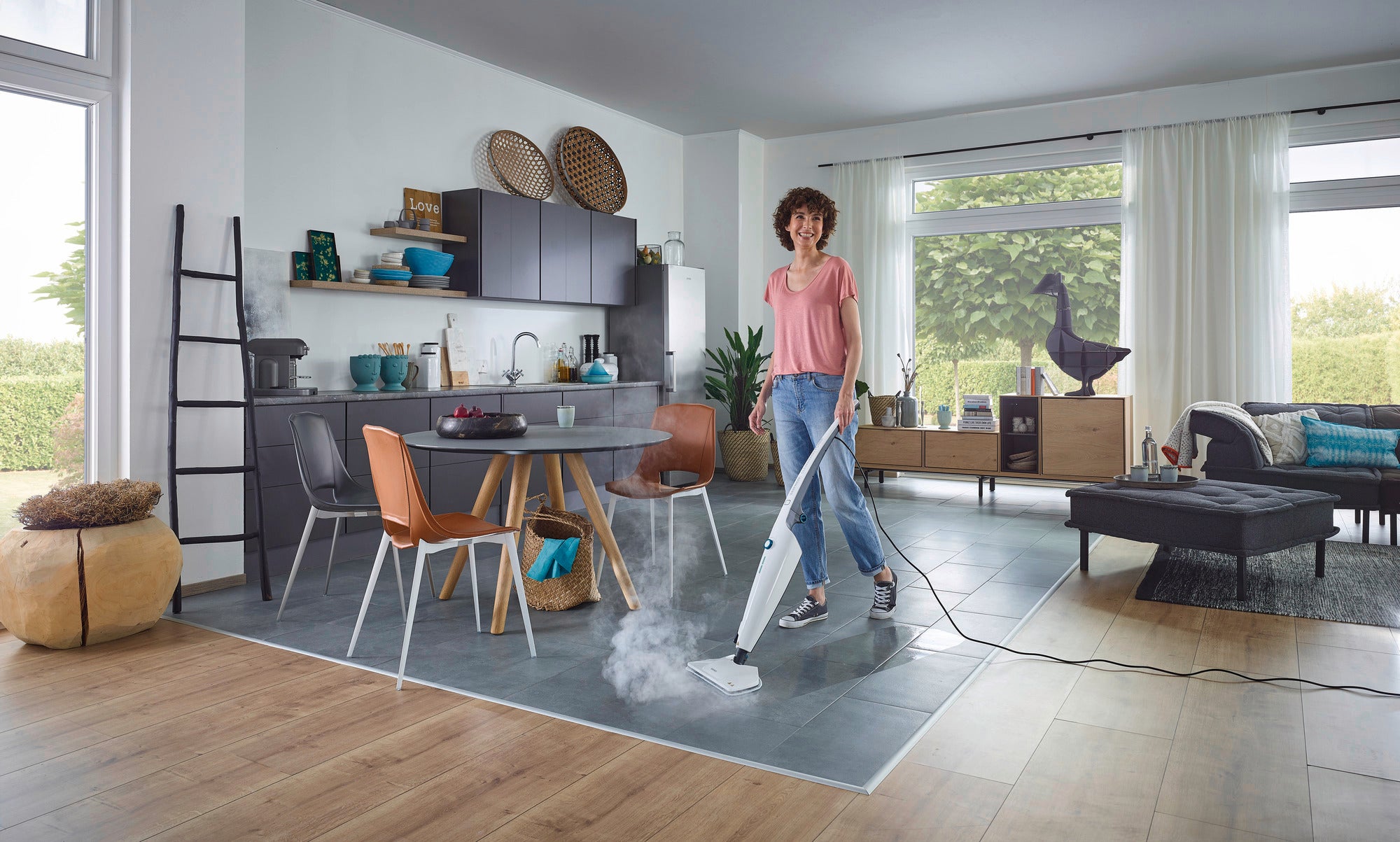 Une femme nettoie le sol d'un salon moderne avec le nettoyeur vapeur CleanTenso Power. La pièce est lumineuse et élégamment meublée, avec de grandes fenêtres et un mobilier moderne.