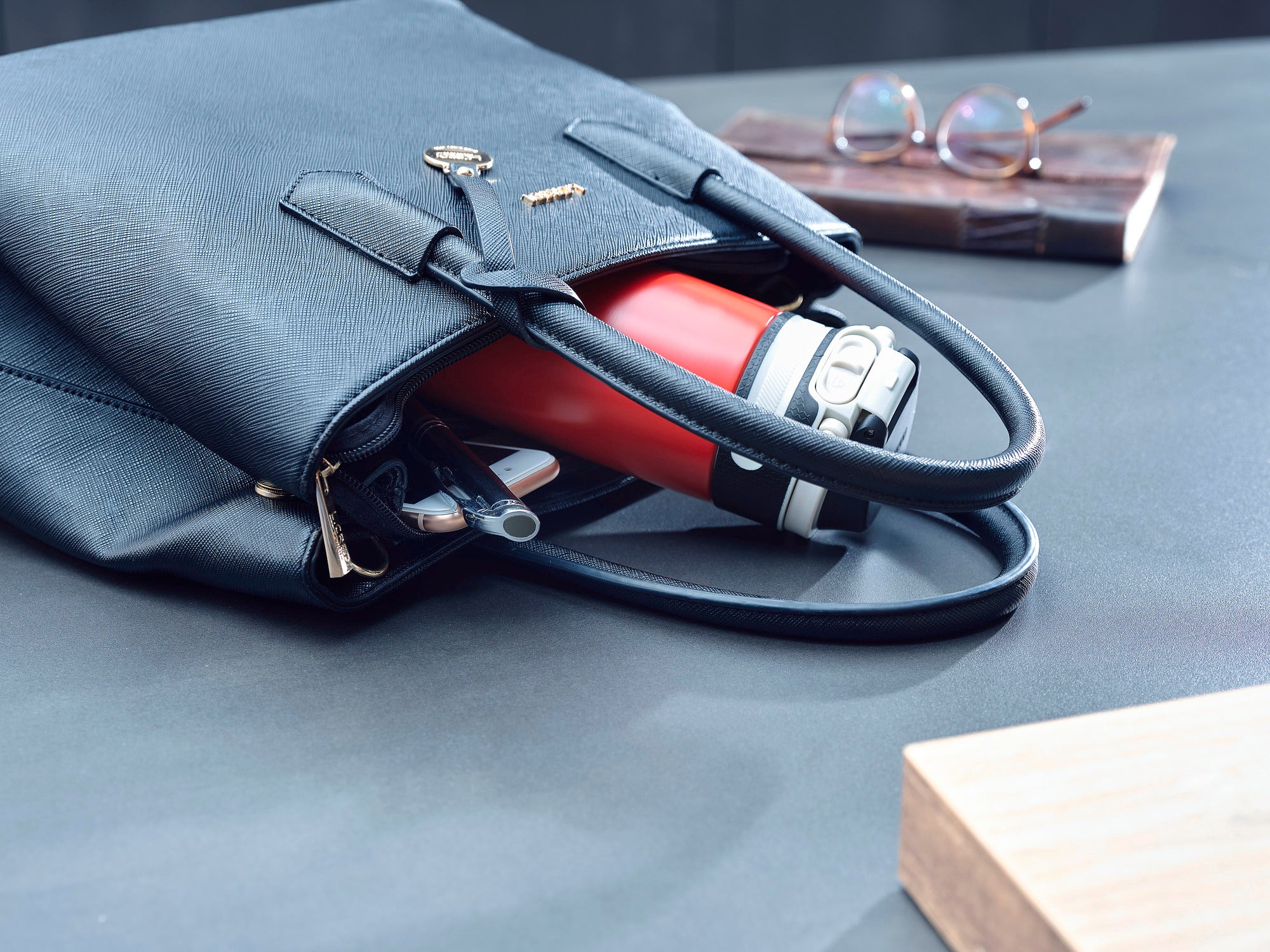 Le mug isotherme Flip 350 ml en rouge est posé dans un sac noir ouvert sur une table. À côté du sac se trouvent des lunettes et un bloc de bois. Idéal pour les déplacements.