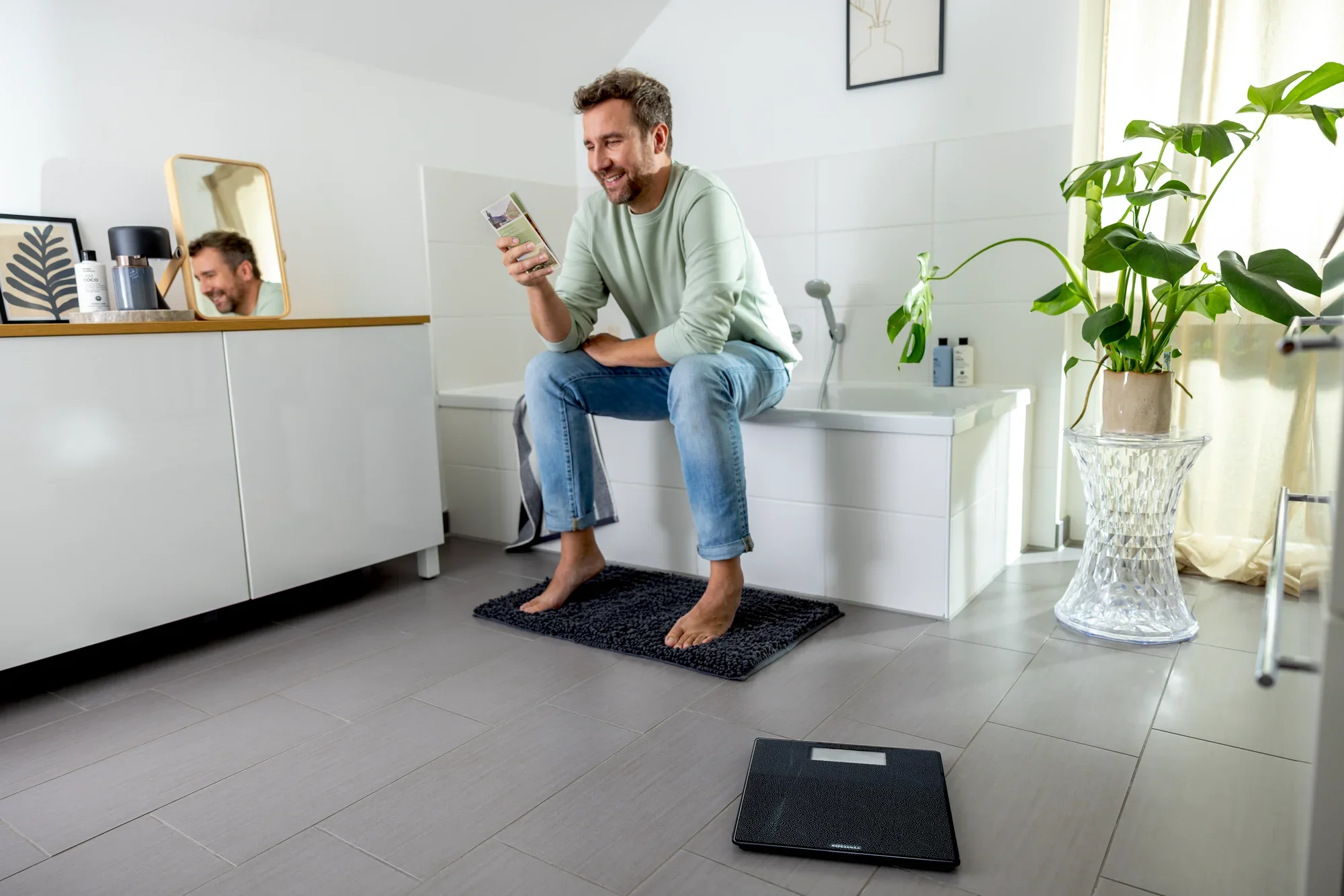 Mężczyzna siedzi na brzegu wanny w łazience i patrzy na swój smartfon. Na podłodze leży cyfrowa waga łazienkowa, model Style Sense Comfort Coral.