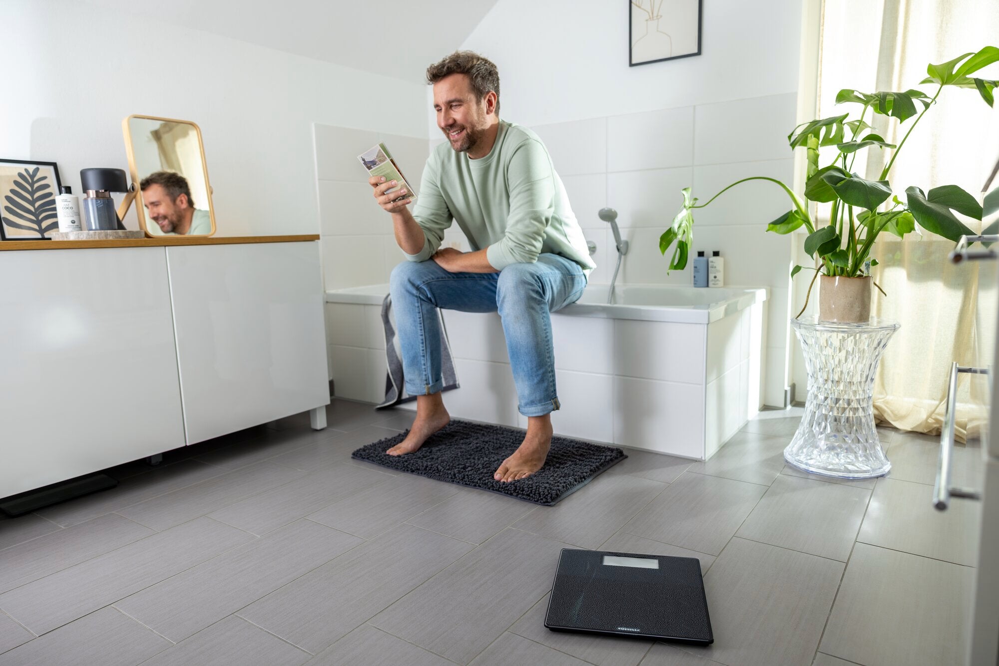 Mężczyzna siedzi na brzegu wanny w łazience i patrzy na swój smartfon. Na podłodze leży cyfrowa waga łazienkowa, model Style Sense Comfort Coral.