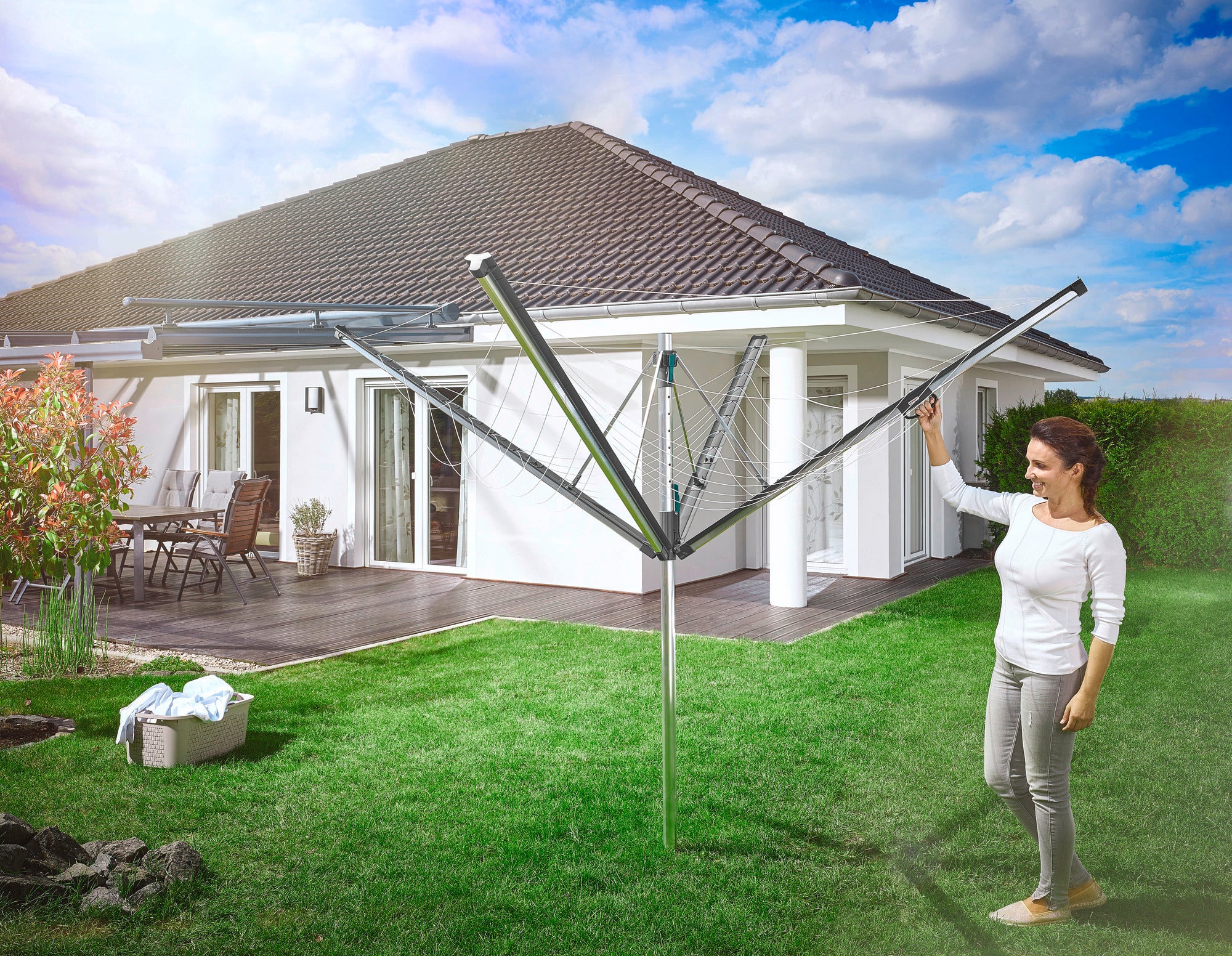 A woman stands in the garden next to an open Linomatic 500 Plus rotary clothes dryer. A modern detached house with a terrace can be seen in the background. The scene shows practical everyday use.