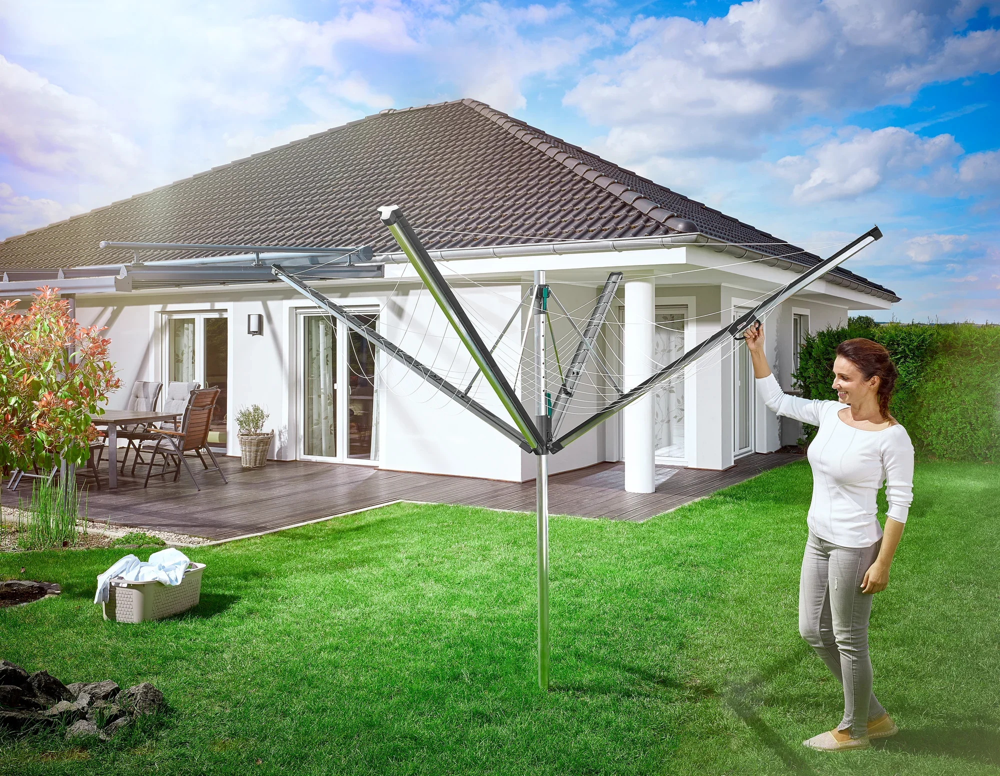 Une femme se tient dans le jardin à côté d'un séchoir à linge Linomatic 500 Plus ouvert. À l'arrière-plan, on aperçoit une maison individuelle moderne avec une terrasse. La scène montre une utilisation pratique au quotidien.