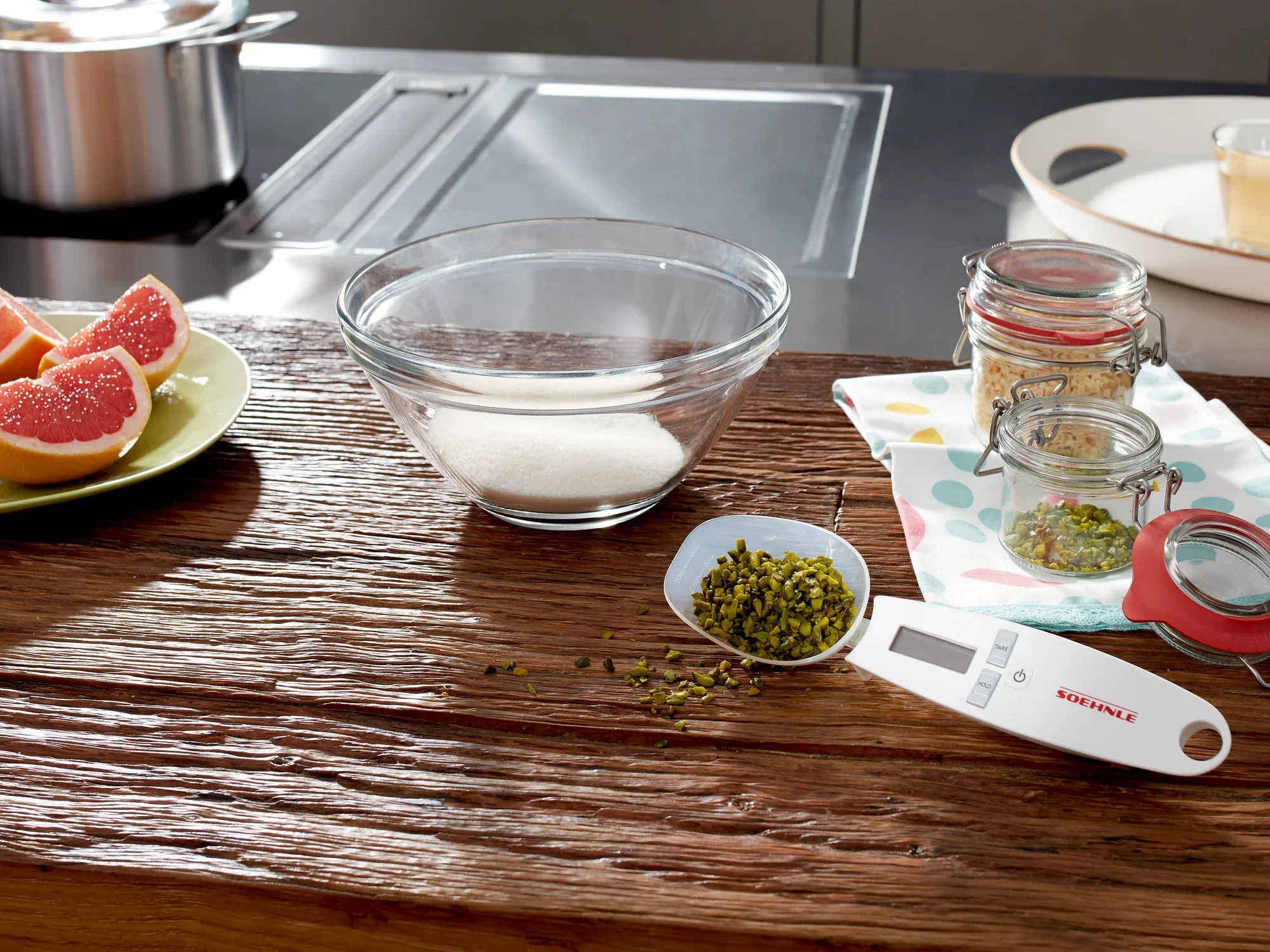 Cyfrowa waga kuchenna Cooking Star leży na rustykalnym drewnianym stole. Obok znajduje się miska z cukrem, słoiki z przyprawami i świeży grejpfrut.